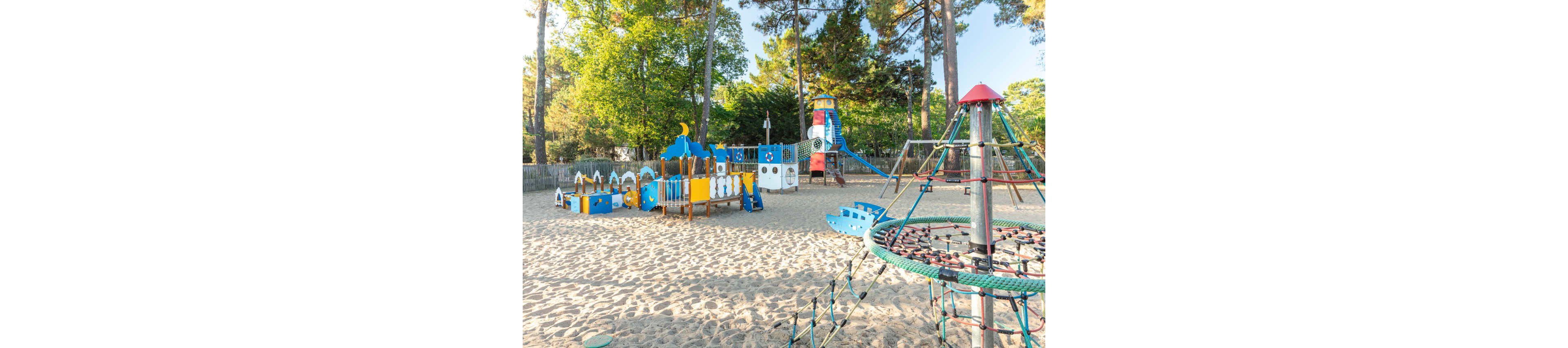 A bright, colorful playground stands on a wide sandy surface surrounded by tall trees. The play structures include a blue and yellow climbing area, slides, and a tower shaped like a lighthouse. In the foreground, a rope climbing frame rises from the sand. The area is empty of children, and soft sunlight filters through the trees, creating a calm outdoor atmosphere.