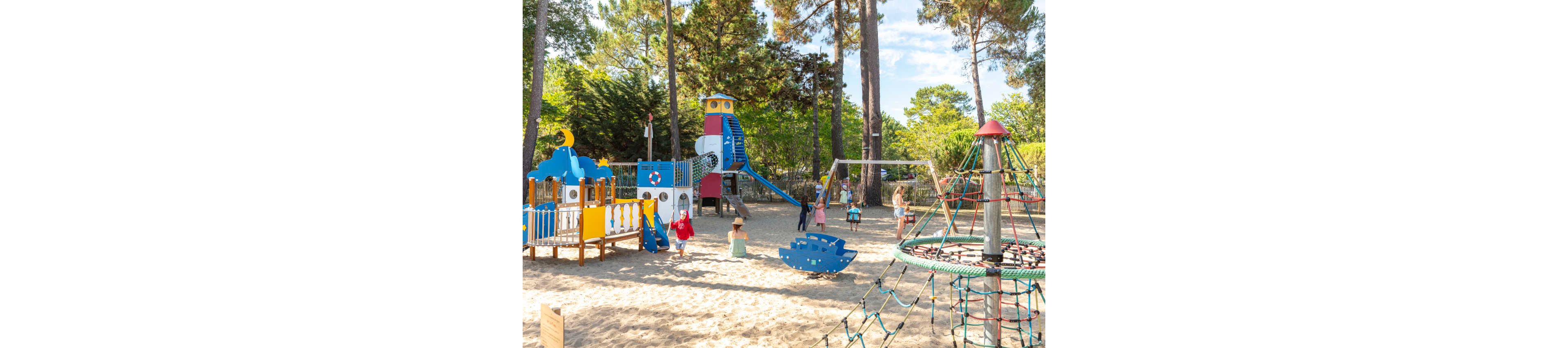Children play in a large outdoor playground surrounded by tall pine trees. The area features a nautical-themed climbing structure shaped like a lighthouse, swings, and a rope climbing pyramid on a sandy surface. Several kids are running, climbing, and swinging while a few adults supervise nearby. The bright colors of the equipment and clear blue sky create a cheerful, summery atmosphere.