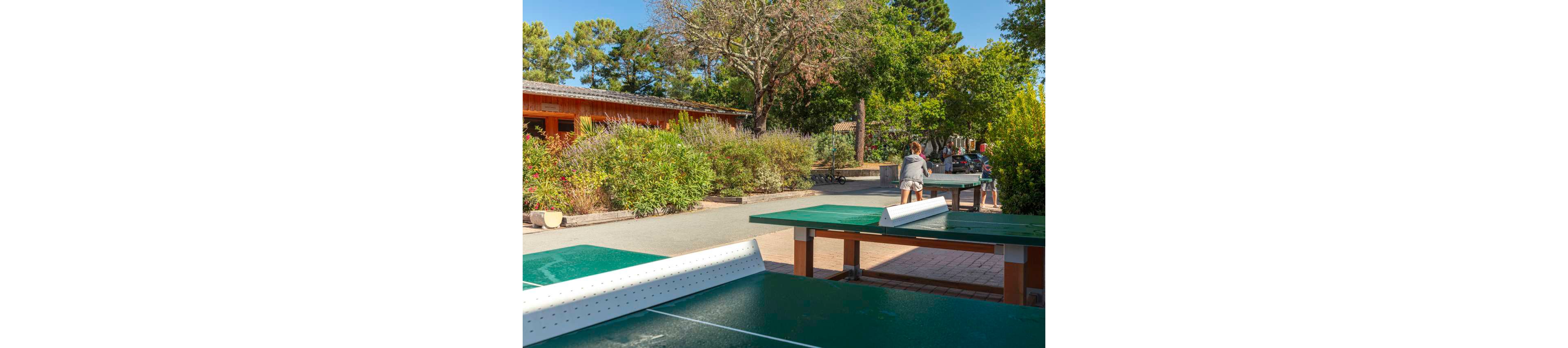 An outdoor recreation area features several green ping pong tables in the foreground. In the background, two people play table tennis near a wooden building surrounded by lush greenery. Trees, shrubs, and bright sunlight create a relaxed, vacation-like atmosphere. The scene suggests a family-friendly leisure space in a park or holiday resort.