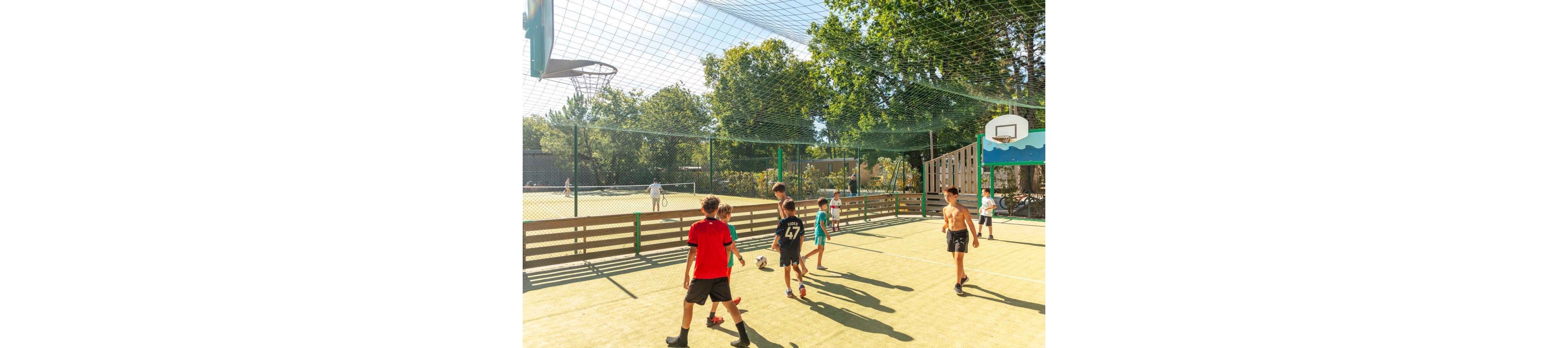 A group of children play soccer on a fenced outdoor court under a protective net. Several boys in sportswear chase the ball while others watch or wait to join. Trees and greenery surround the court, and a basketball hoop is visible in the background. The bright sunlight and clear sky create a lively, energetic atmosphere.