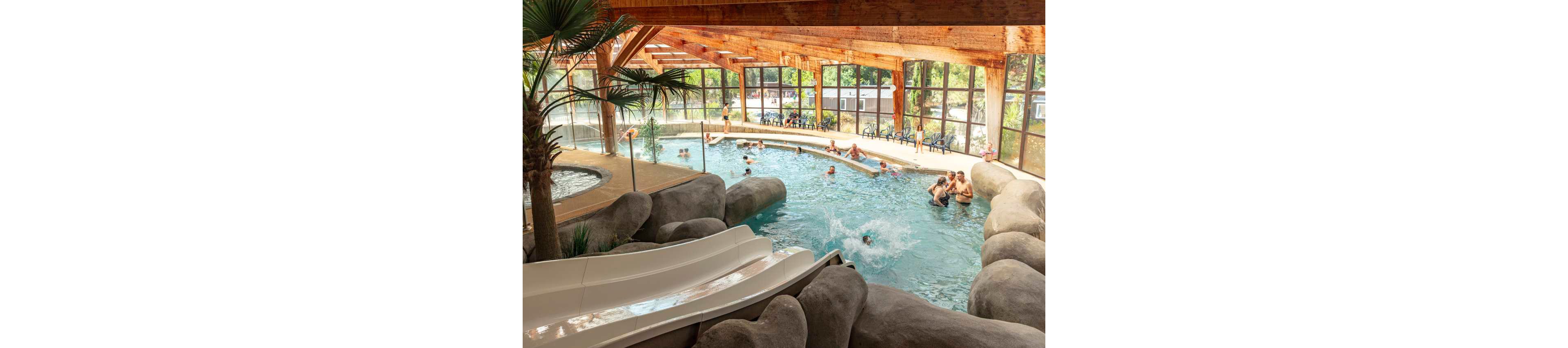 An indoor leisure pool features families and children enjoying the water under a wooden beamed roof. A white waterslide curves between artificial rock formations into the main swimming area. Large windows surround the pool, revealing green trees and parked caravans outside. People relax on poolside chairs while others swim, splash, and play in the warm, bright environment.