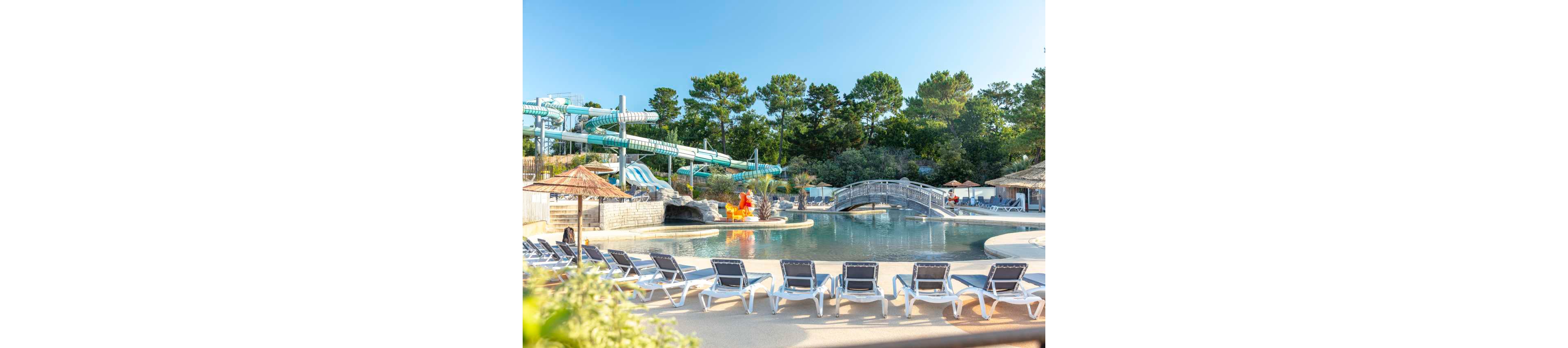 An outdoor water park features a calm lagoon-style swimming pool surrounded by empty lounge chairs. A curved footbridge crosses the pool, leading toward a cluster of tall water slides in the background. Trees and greenery frame the scene under a clear blue sky, suggesting a warm summer day. The overall atmosphere is relaxed and inviting, ideal for family vacations or resort leisure.