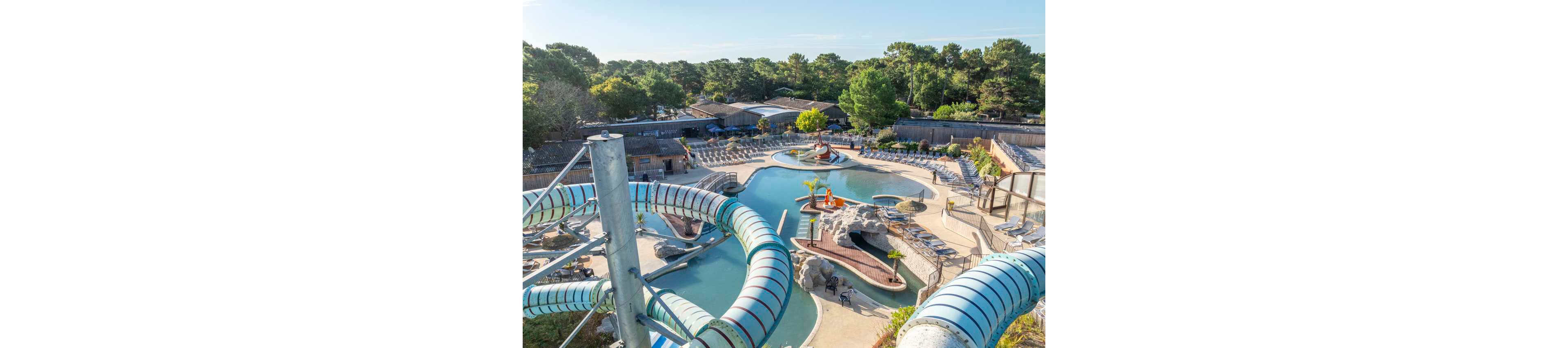 An outdoor water park features a large central pool surrounded by winding tube water slides. Sun loungers and umbrellas line the poolside terraces, creating a relaxing resort atmosphere. Decorative rocks, small bridges, and a children’s play area add visual interest to the aquatic space. Dense green trees and cabins in the background suggest a holiday park or campground setting.