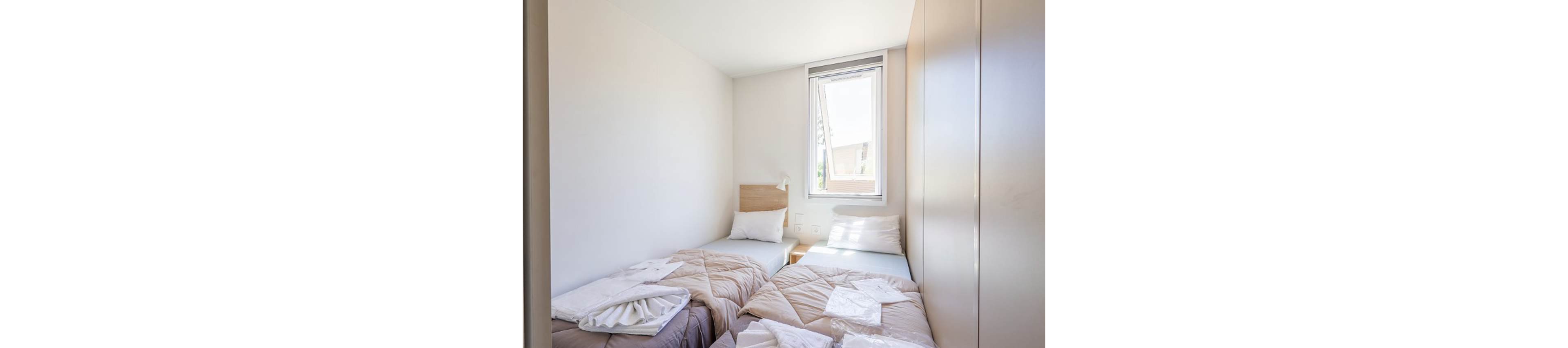 Compact twin bedroom with two single beds is shown in bright natural light. The beds are neatly made with beige duvets, white sheets, and folded towels placed on top. A tall wardrobe stands along one wall, and a small headboard with a reading light is visible. A single window lets in daylight and offers a partial view of neighboring buildings.