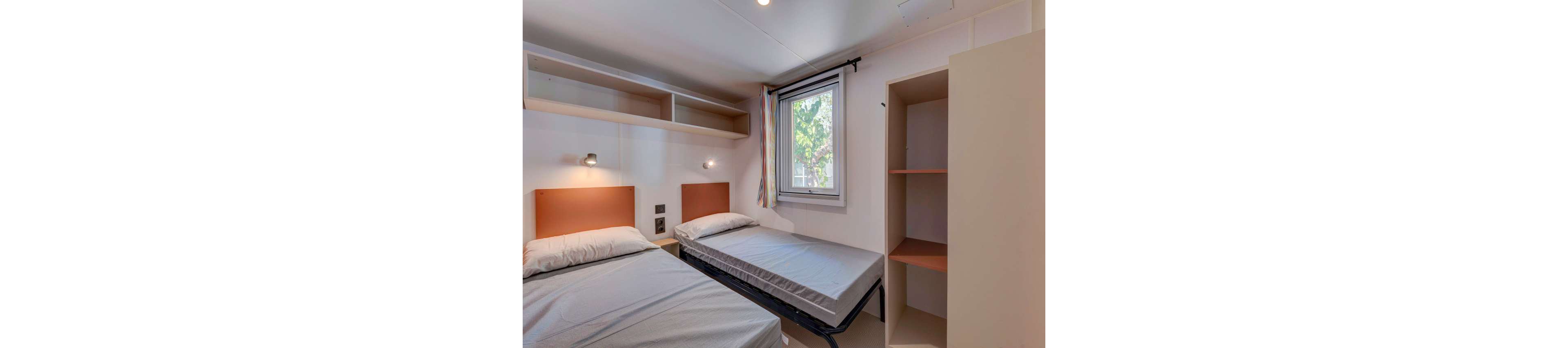 Small bedroom featuring two single beds with simple headboards and neutral bedding. One bed is slightly pulled out, suggesting a flexible sleeping arrangement. The room includes open wall shelves and an empty open wardrobe for storage. A window with colorful curtains lets in natural light, brightening the minimalist interior.