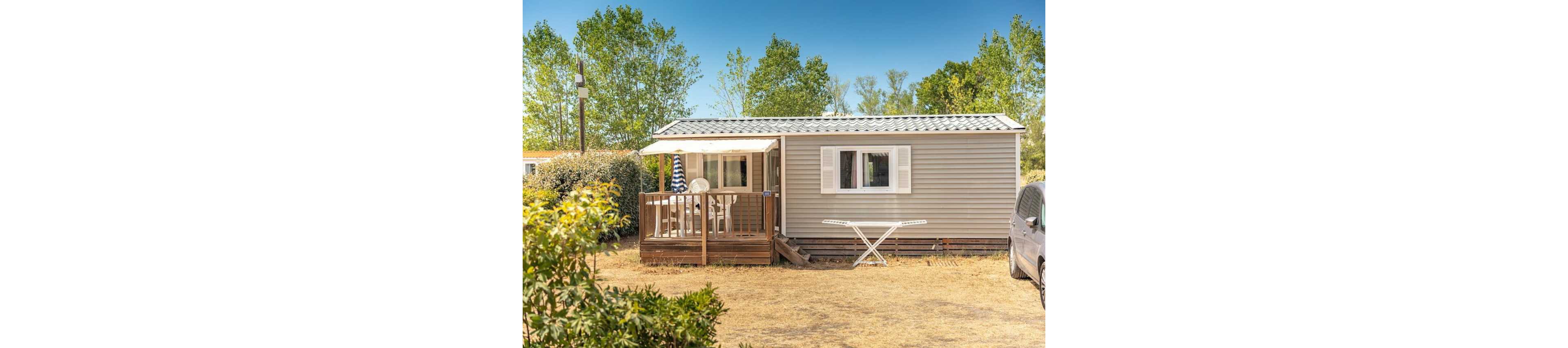 A compact mobile home stands in a dry, sunlit yard with trees in the background. The front features a wooden deck furnished with a table, chairs, and a striped patio umbrella. An empty white drying rack is set up on the ground in front of the home, and part of a parked vehicle is visible on the right side. The scene suggests a relaxed vacation or campsite setting in warm weather.
