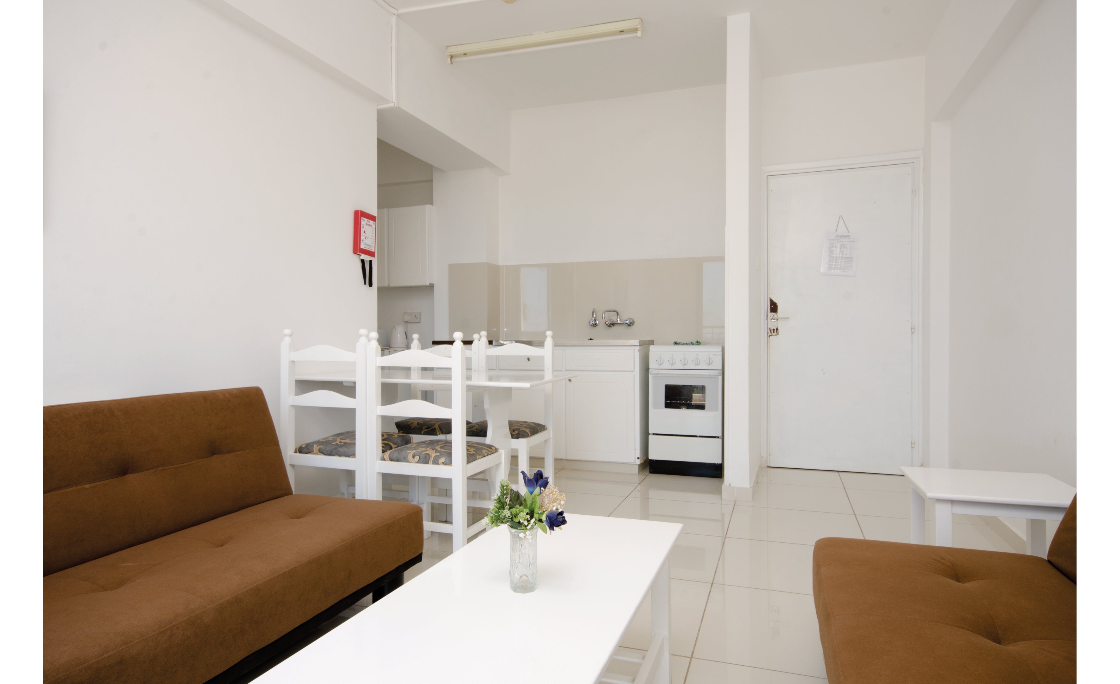 A compact apartment living space featuring a small kitchenette and dining area. The setting includes brown sofas, a white dining table with chairs, and a vase with flowers on the coffee table. The room is bright with white walls and tiled flooring, creating a clean and minimalistic look. A fire extinguisher is visible on the wall.