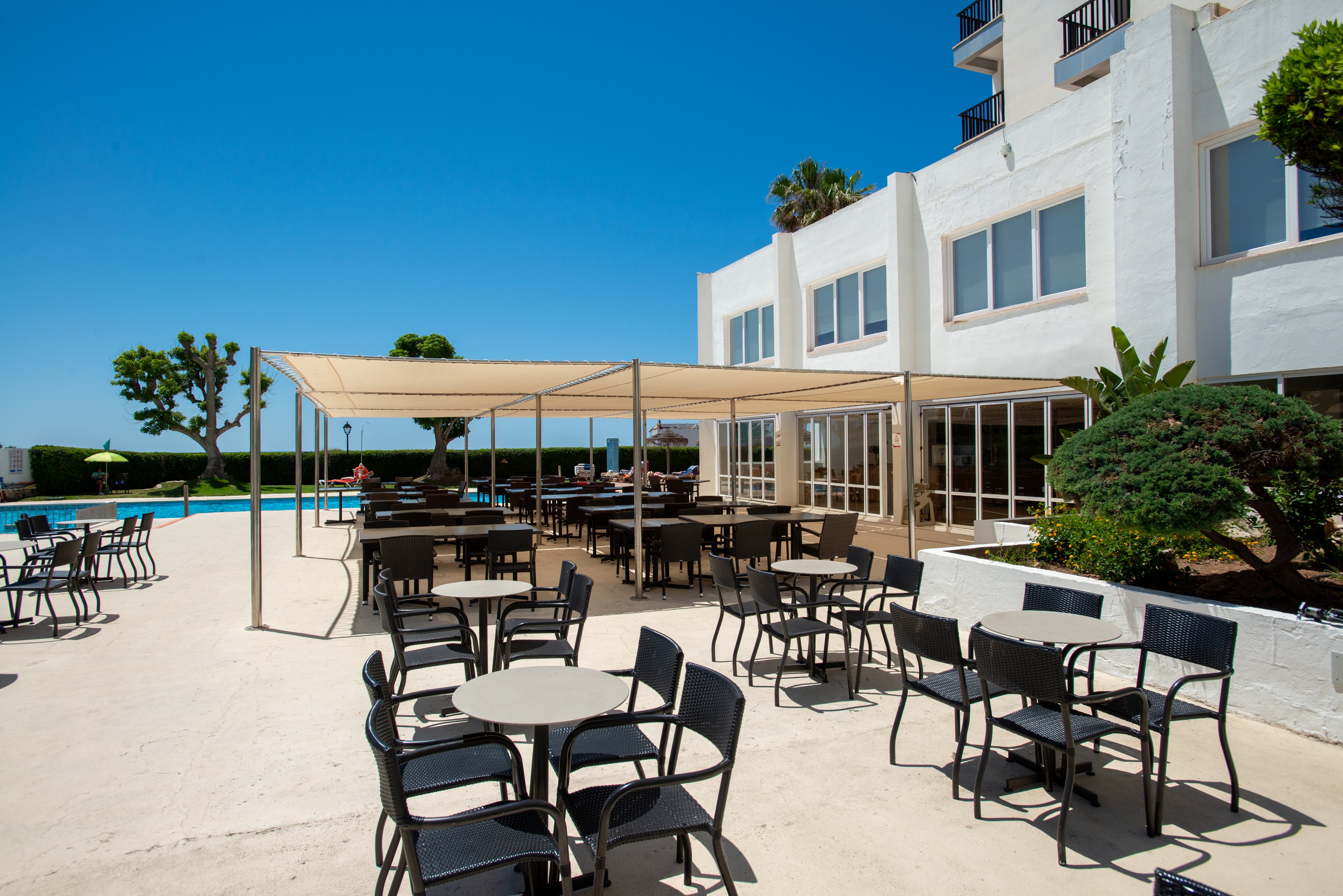The image shows a bright outdoor terrace area at a hotel or resort with numerous tables and chairs arranged for guests. A large canopy provides shaded seating near the building’s glass doors and windows. In the background, a swimming pool and manicured trees are visible under a clear blue sky. The overall scene suggests a relaxed, vacation atmosphere in a warm, sunny climate.