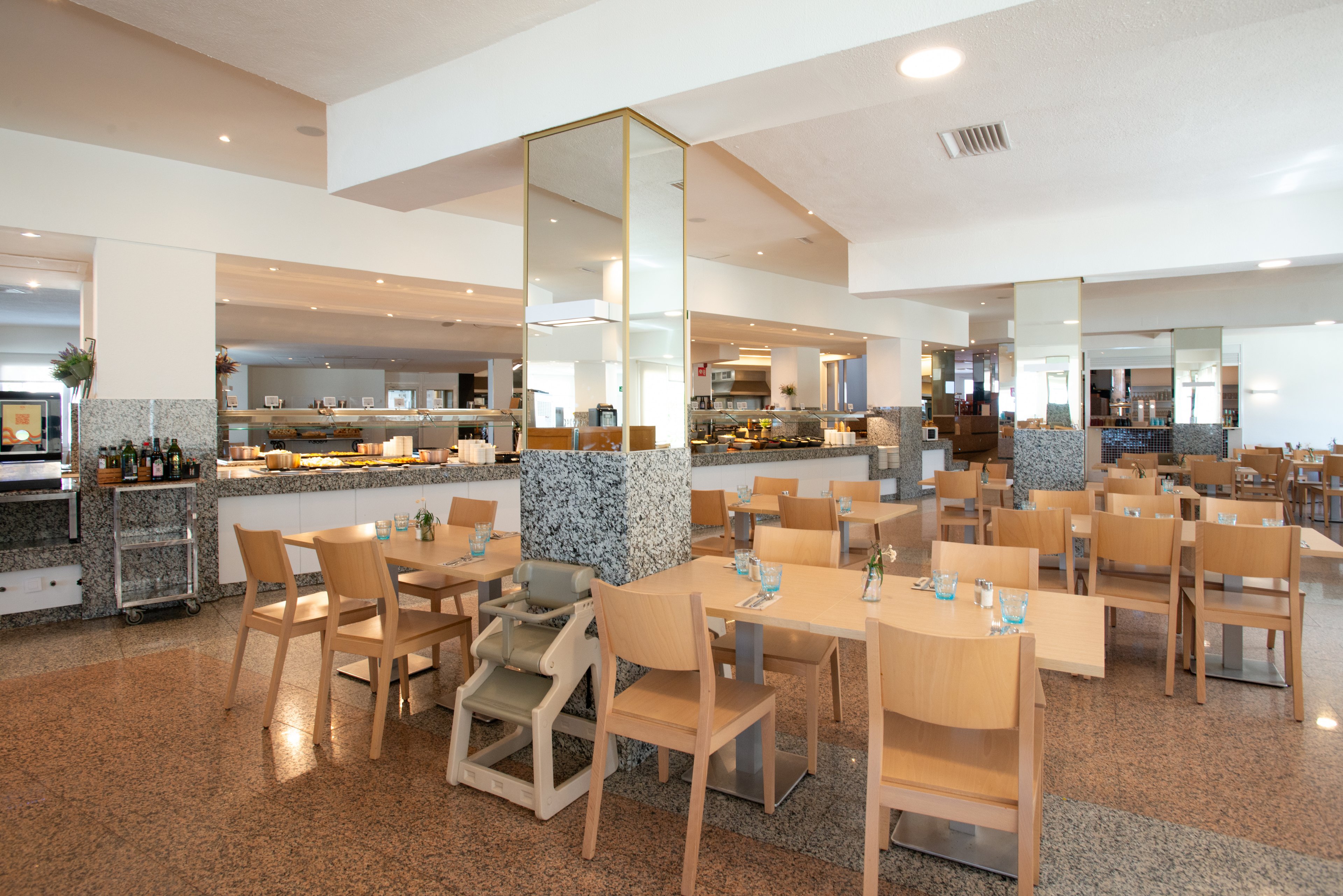 Spacious restaurant interior featuring light wooden tables and chairs arranged neatly across a polished floor. A buffet counter with various dishes and beverages is visible in the background, suggesting a self-service dining experience. Large mirrored columns and recessed ceiling lights create an open, contemporary atmosphere. Table settings with glasses and cutlery are prepared, and a wooden high chair indicates family-friendly seating.