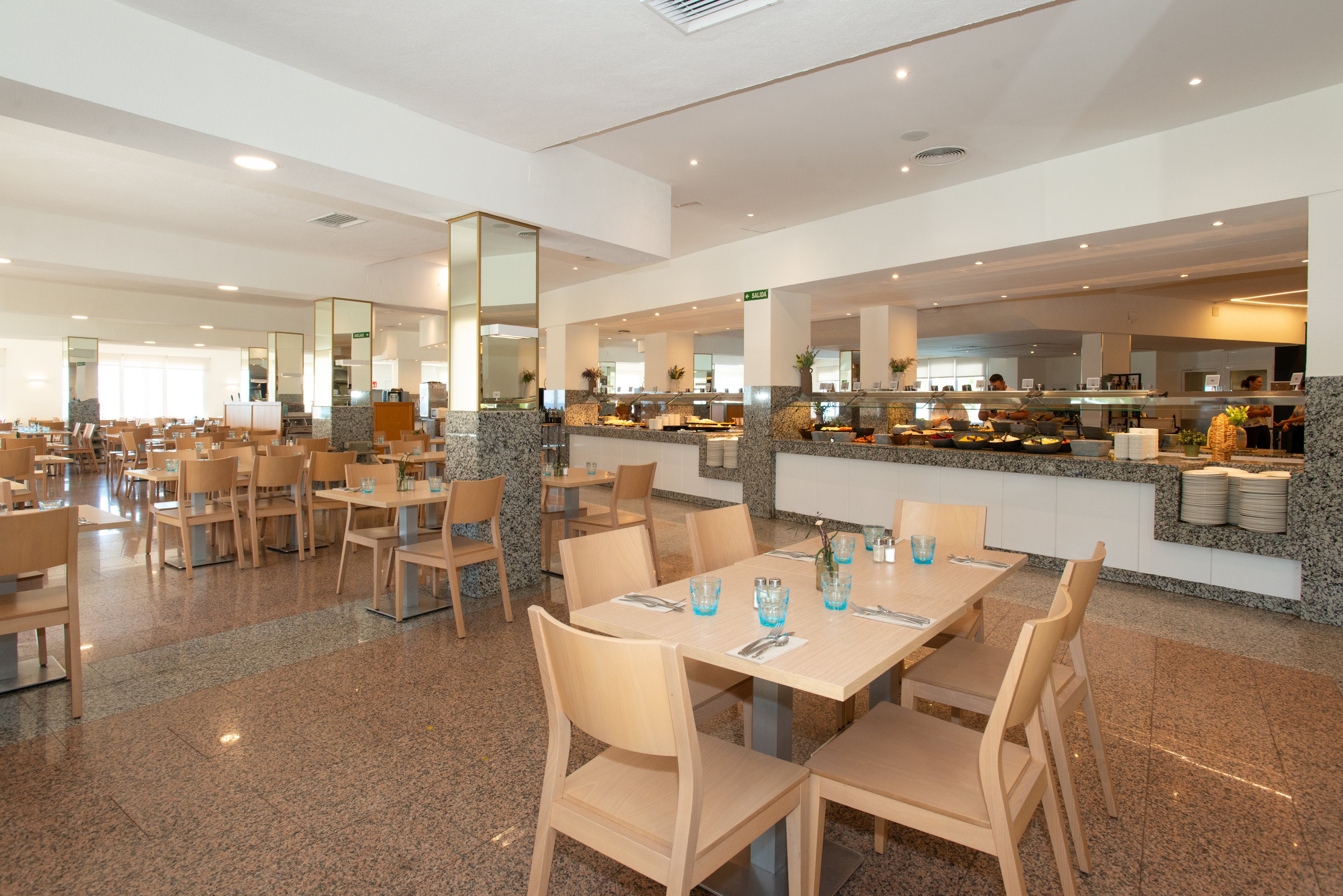 Spacious self-service restaurant interior with light wooden tables and chairs arranged neatly across a polished floor. The buffet counter in the background displays various dishes and serving stations under warm lighting. Glasses, cutlery, and napkins are set on the tables, ready for guests. Large mirrors and wide open walkways contribute to an airy, contemporary atmosphere suitable for casual dining.