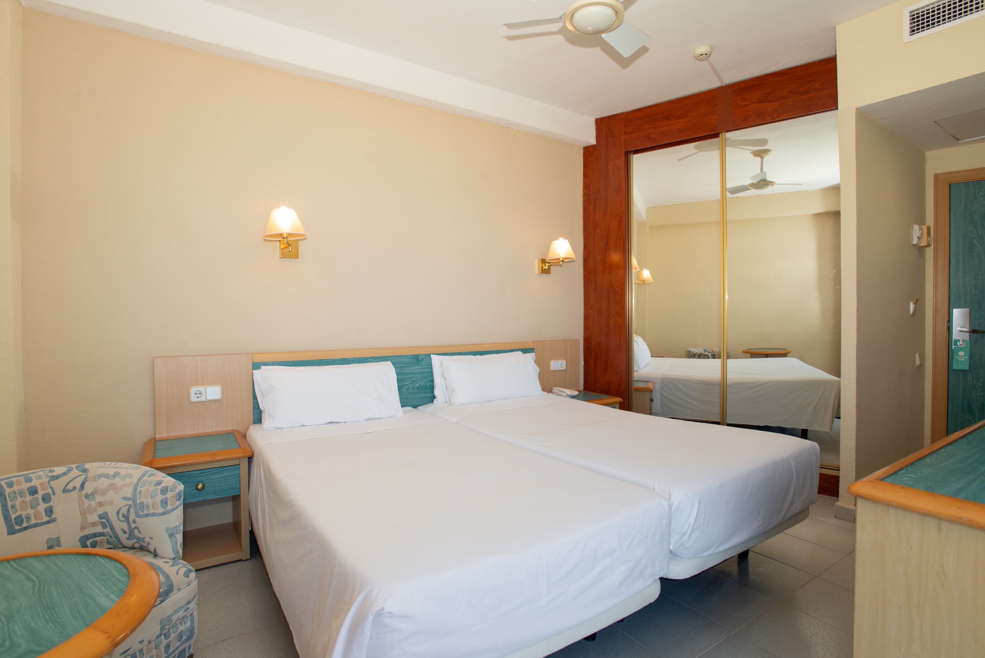 A clean hotel room features two twin beds pushed together with white linens and teal accents. The space includes bedside tables, wall-mounted lamps, and a patterned armchair. A large mirrored wardrobe reflects the room, adding depth and brightness. Neutral walls and simple decor create a calm, minimalist atmosphere suitable for guests.