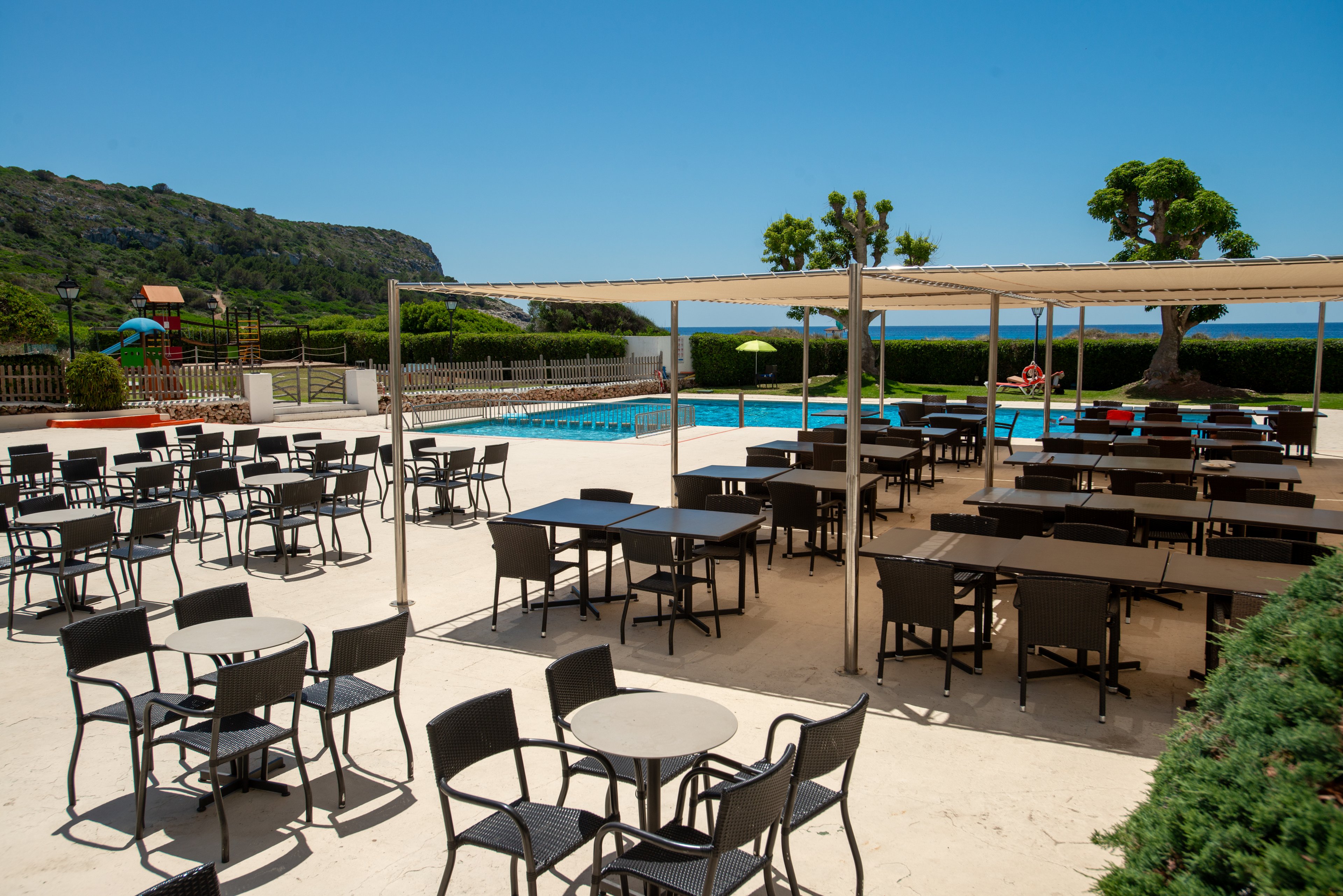 An empty outdoor terrace is filled with black tables and chairs arranged in neat rows. The area is covered by pergola-style canopies, providing shade near a bright blue swimming pool. Lush green hedges, trees, and distant hills frame the scene under a clear, sunny sky. The setting suggests a tranquil resort or hotel environment ready for guests.