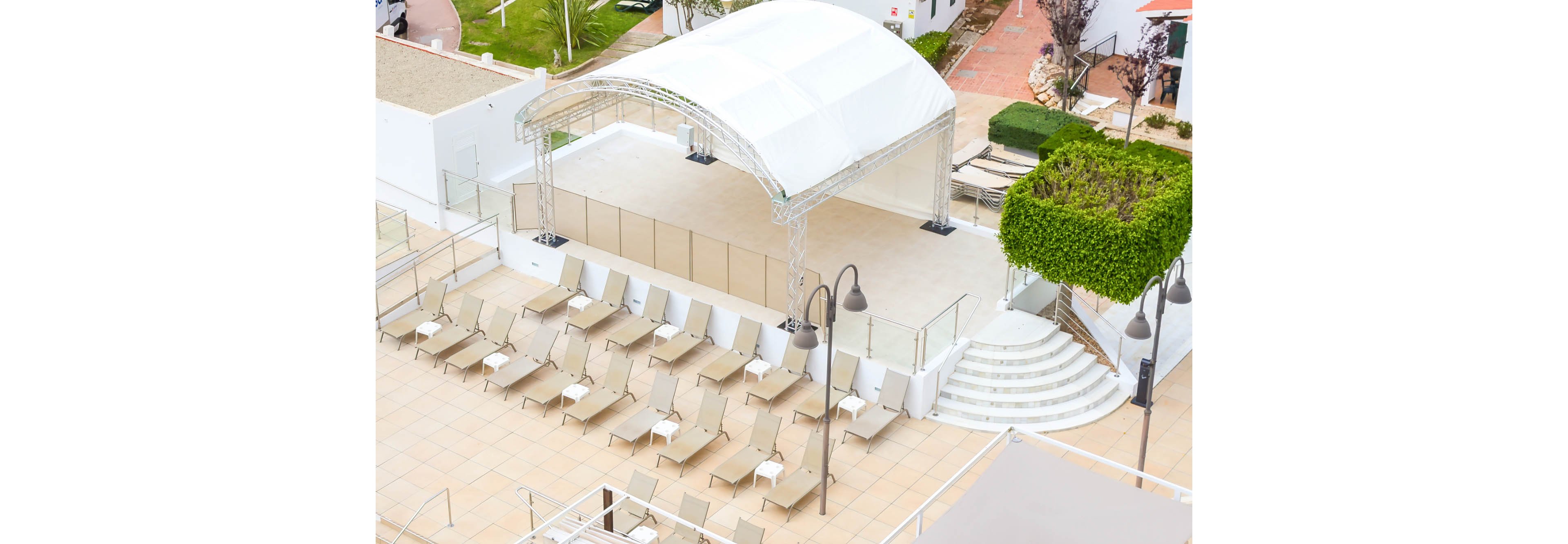 An elevated view shows an empty resort sun deck lined with rows of beige lounge chairs. A white canopy stage structure stands at one end, framed by glass railings and metal supports. Curved white steps lead up from a tiled courtyard, with manicured green shrubs and pathways in the background. The bright, neutral color palette and absence of people create a calm, orderly atmosphere.