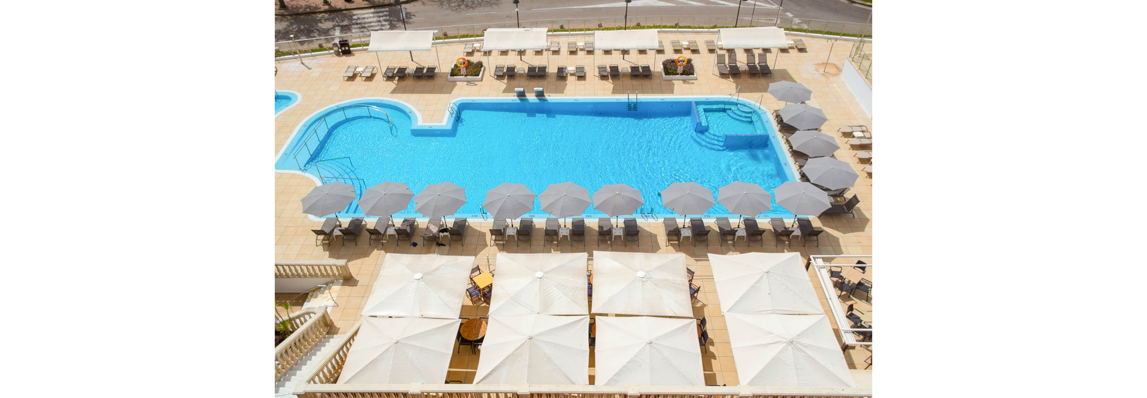 An elevated view shows a bright blue hotel swimming pool surrounded by sun loungers and umbrellas. The pool has a distinctive rounded shape with steps leading into the shallow end. Rows of neatly arranged parasols and shaded seating areas line the tiled pool deck. The scene appears calm and empty, suggesting a quiet resort atmosphere on a sunny day.