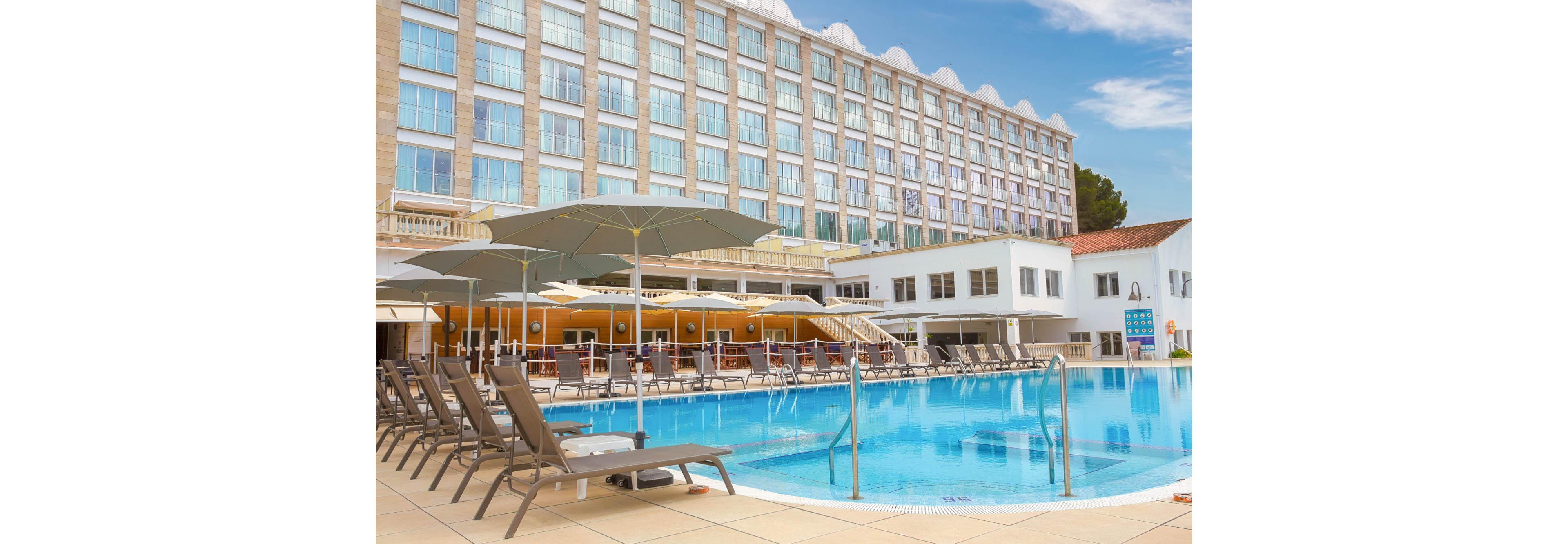 A large outdoor swimming pool sits in front of a modern multi-story hotel building. Numerous sun loungers and umbrellas line the pool deck, creating a relaxing resort atmosphere. The hotel façade features many identical windows, while a poolside bar or terrace area is visible under a shaded structure. Clear blue water and a bright sky emphasize a sunny vacation setting.