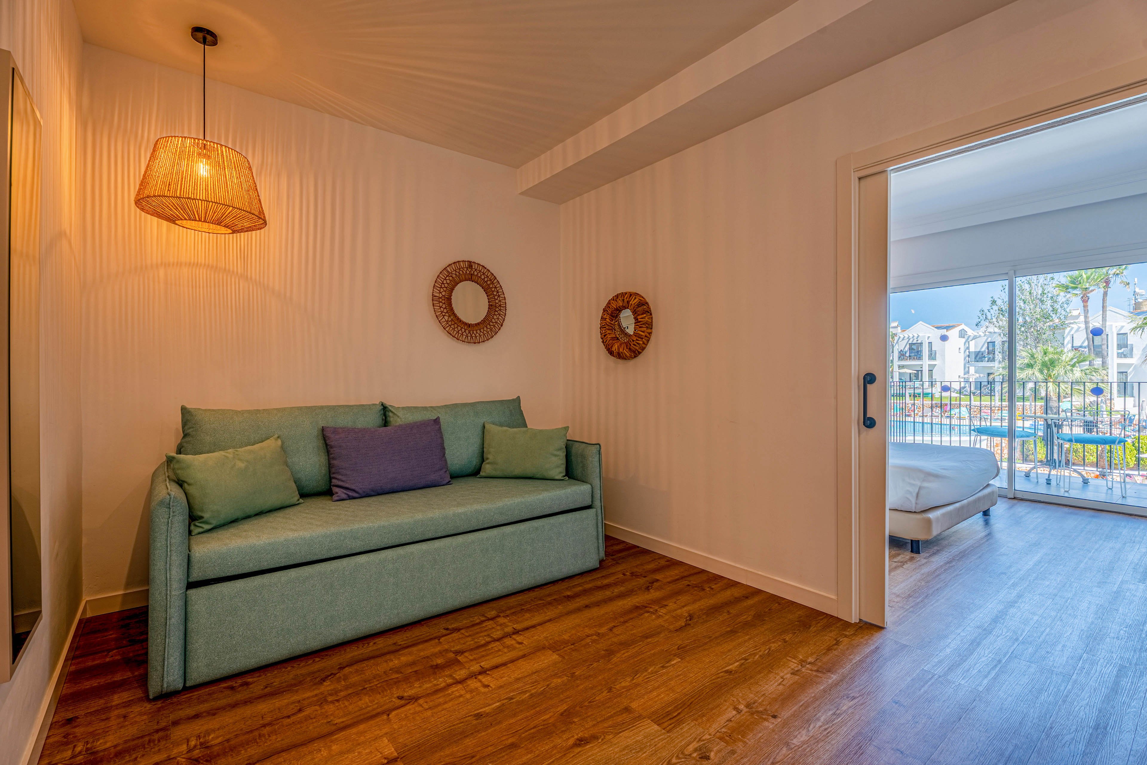 A small sitting room features a teal sofa with colorful cushions against a textured white wall. A warm pendant light hangs from the ceiling, casting a soft glow over the wooden floor. Two round decorative wall pieces add visual interest beside a large mirror. Through the open sliding door, a bright bedroom and balcony overlook a residential complex and greenery.