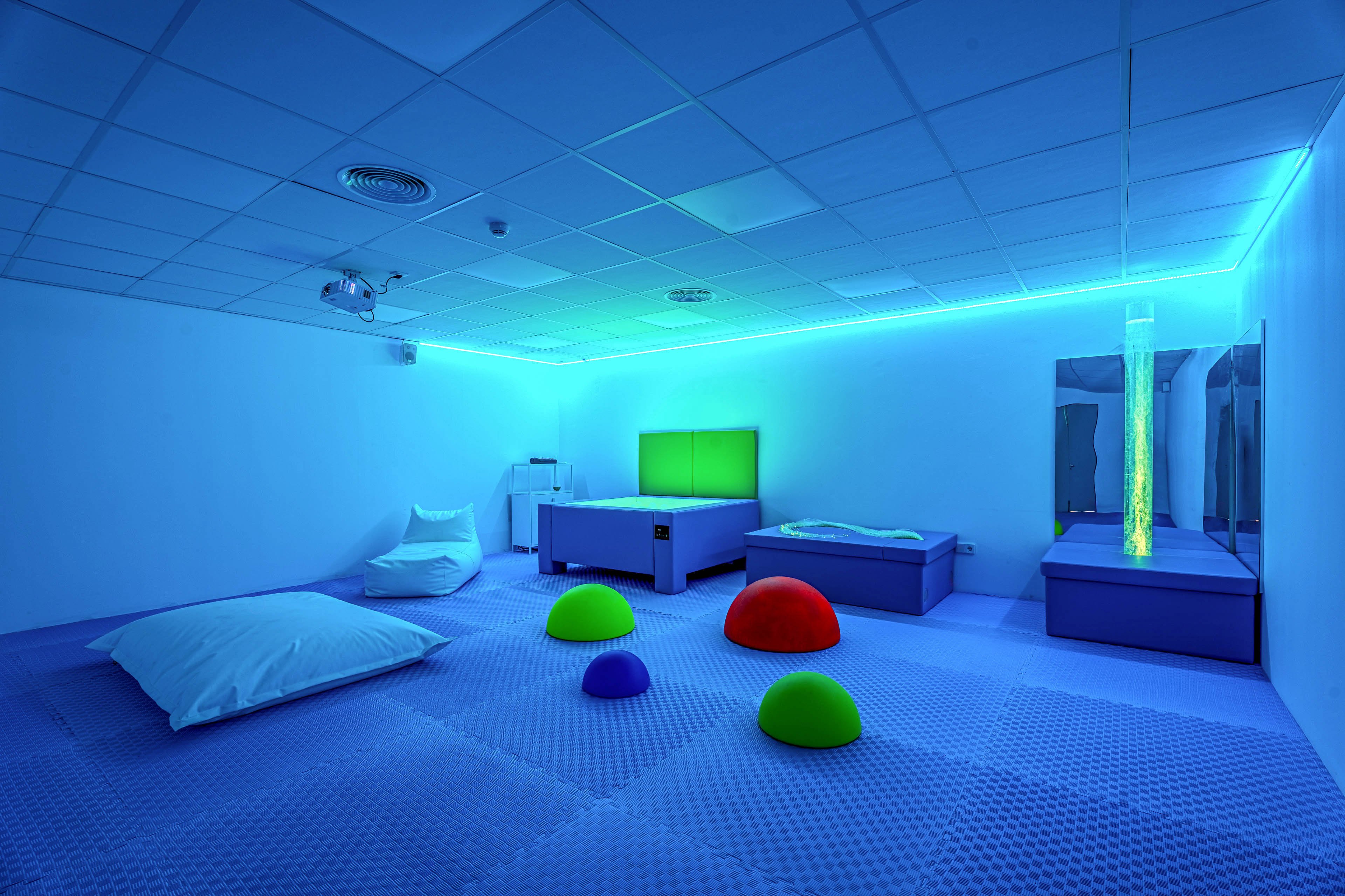 A modern sensory therapy room is illuminated with blue ambient lighting and accent strips along the ceiling and walls. The space features soft padded flooring, a low bed, cushioned bench, and large floor pillow for comfort and safety. Colorful dome-shaped play elements in green, blue, and red add visual stimulation and tactile interaction. A vertical light column and mirrored surfaces enhance the calming, immersive atmosphere of the room.