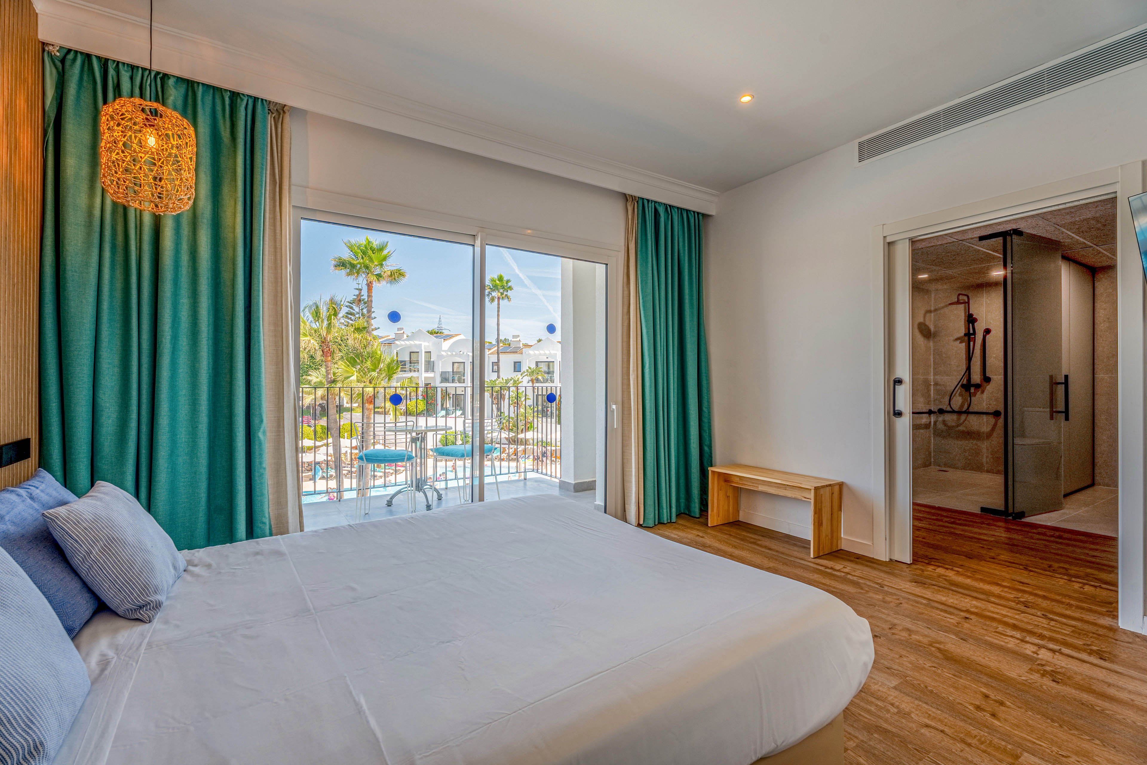 A modern hotel bedroom features a large neatly made bed facing a balcony with glass doors. Turquoise curtains frame the view of palm trees and white resort buildings outside. Warm wooden flooring and a woven pendant light add a cozy, natural touch to the space. A sliding glass door reveals an adjacent bathroom with a spacious walk-in shower.