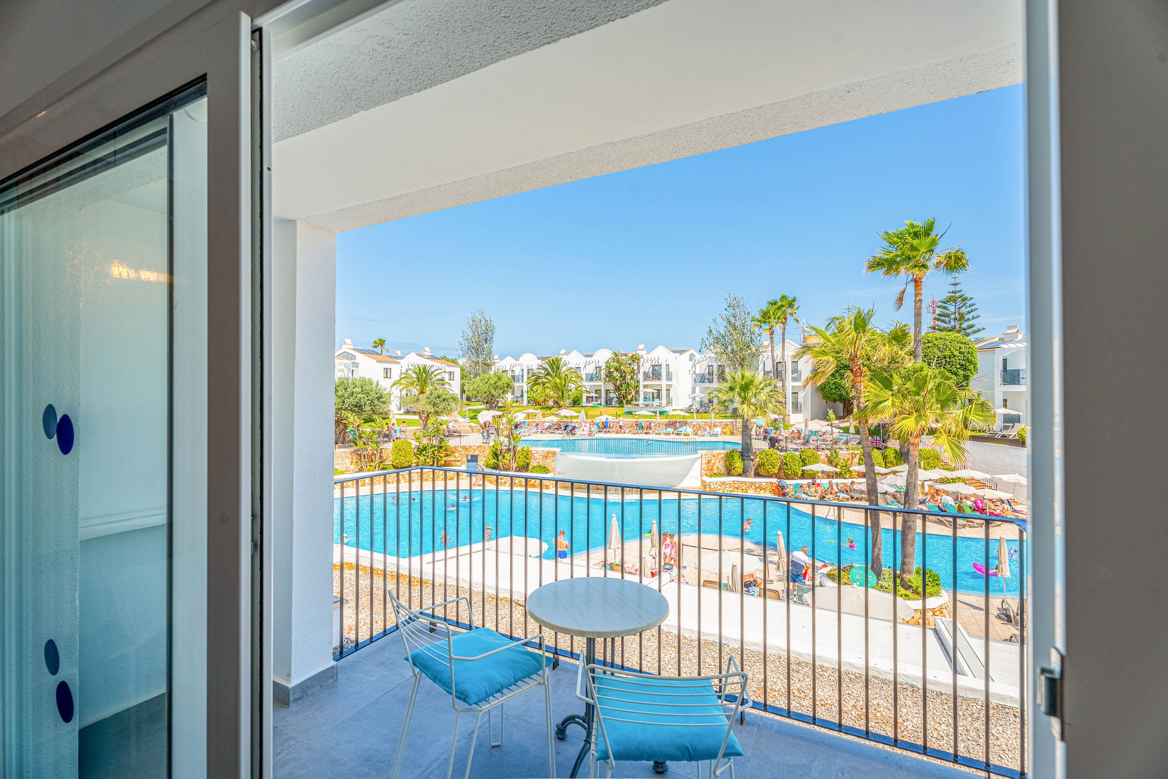 A small balcony with a round table and two blue chairs overlooks a large resort swimming pool. The scene includes sun loungers, umbrellas, and landscaped gardens with palm trees. White low-rise buildings surround the pool under a clear blue sky. The bright colors and calm water create a relaxed vacation atmosphere.