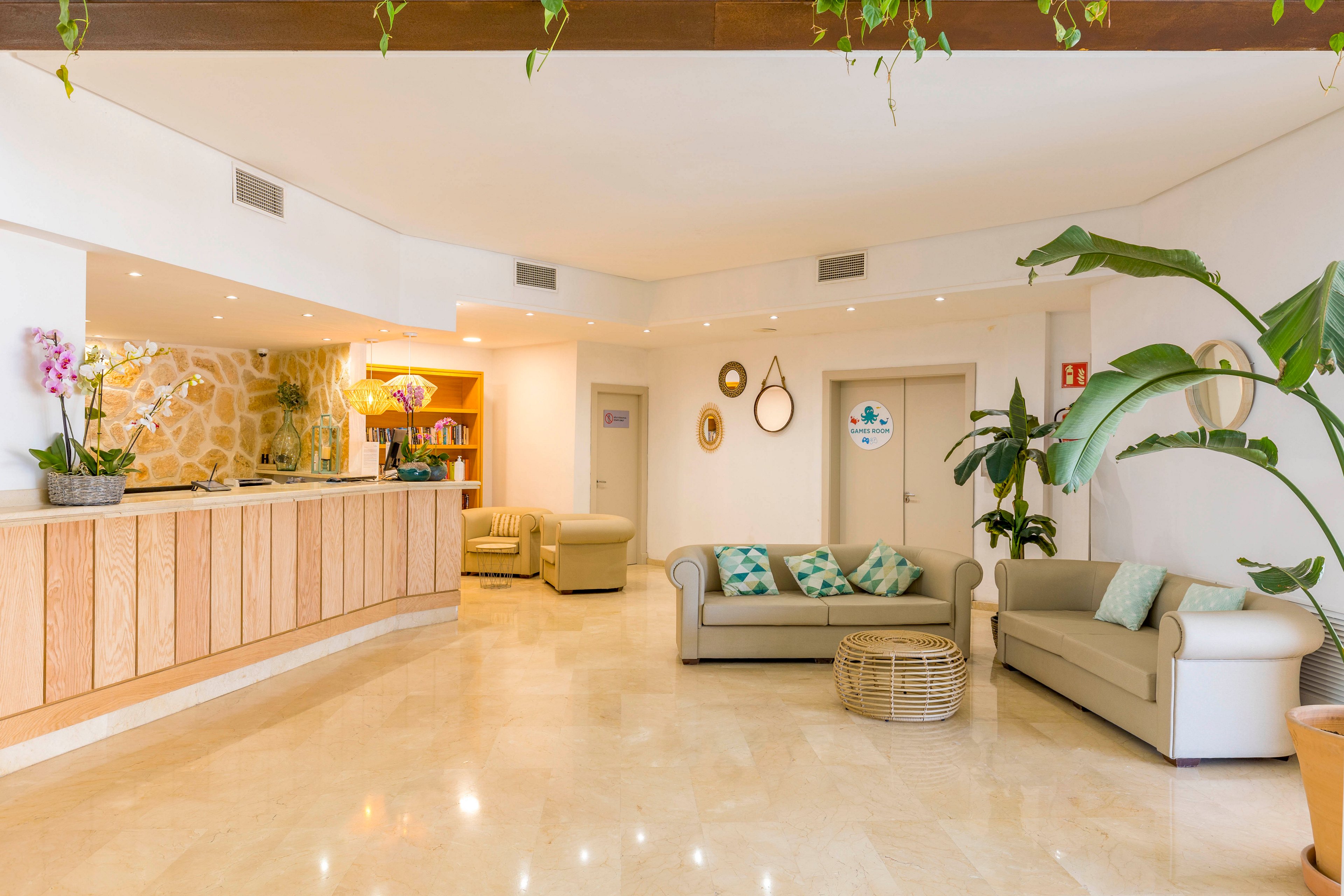A spacious, modern hotel lobby features a light wood reception desk on the left and comfortable sofas with patterned cushions on the right. The room is decorated with indoor plants, wall mirrors, and soft lighting that creates a welcoming atmosphere. Neutral cream flooring and walls are accented by pastel colors and natural textures, including a wicker coffee table. Doors and signage in the background suggest access to additional guest facilities.