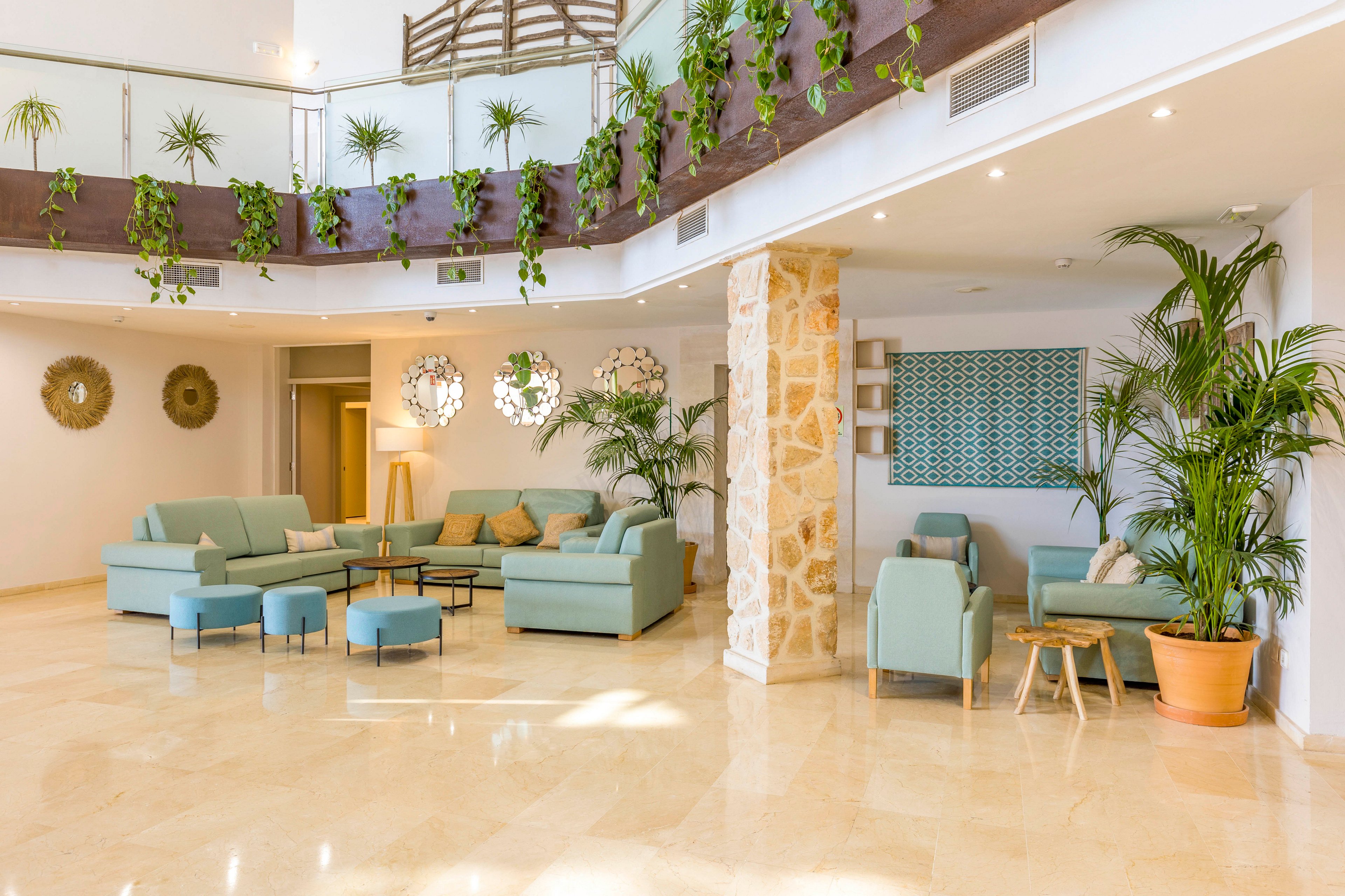Spacious contemporary lobby interior featuring pastel sofas and ottomans arranged for relaxed seating. Large potted plants and hanging greenery decorate the upper balcony and corners, adding a fresh natural touch. Warm stone column, wall art, and decorative light fixtures create a stylish, welcoming atmosphere. The polished floor and white walls enhance the airy, sunlit feel of the space.