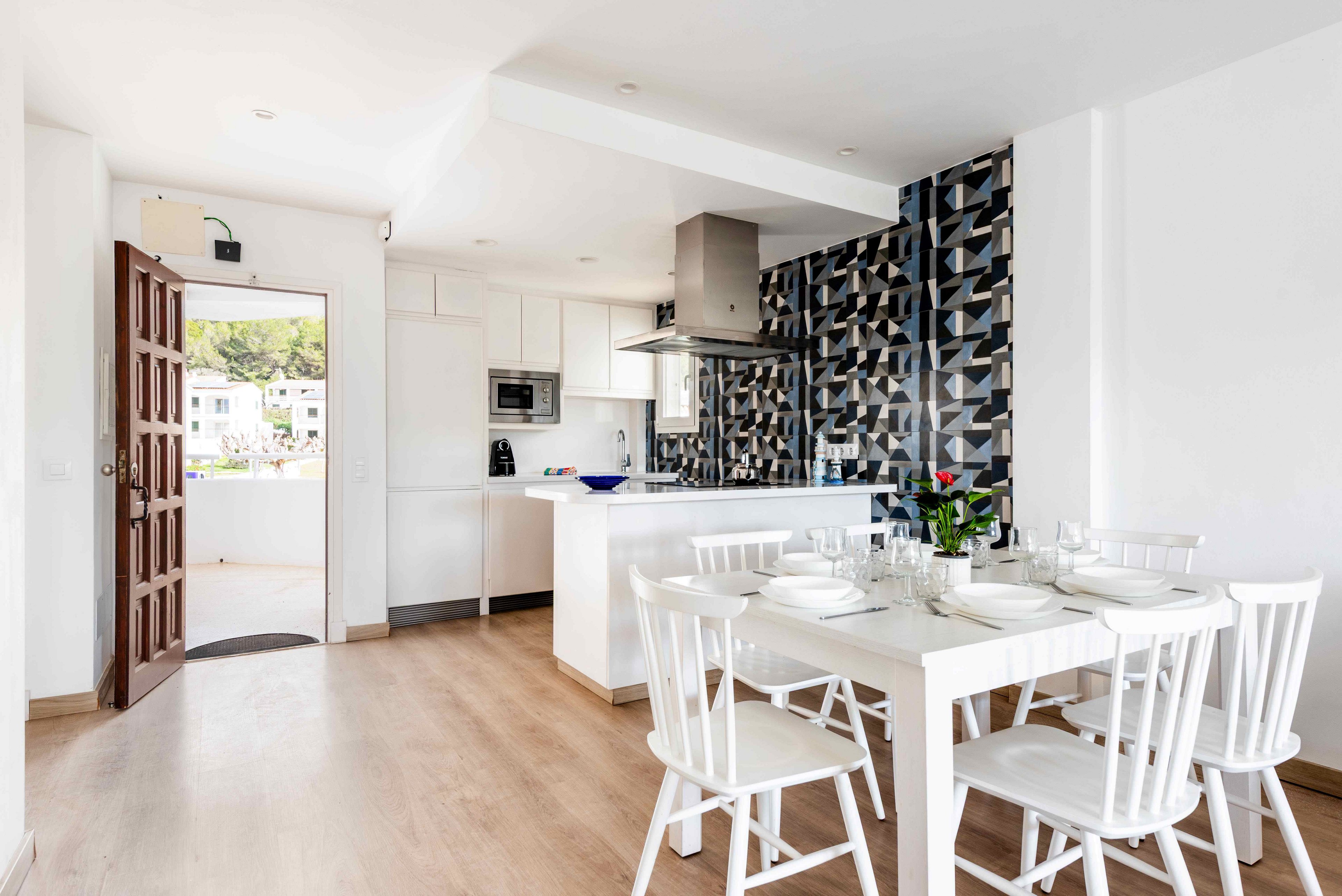 A bright, modern kitchen and dining space features white cabinetry and a striking geometric tile backsplash. The dining table is set for four with white chairs and tableware, and a small plant centerpiece adds a touch of greenery. Natural light fills the room through an open door, highlighting the clean, minimalist decor and light wood flooring. No visible text or numbers are present in the image.
