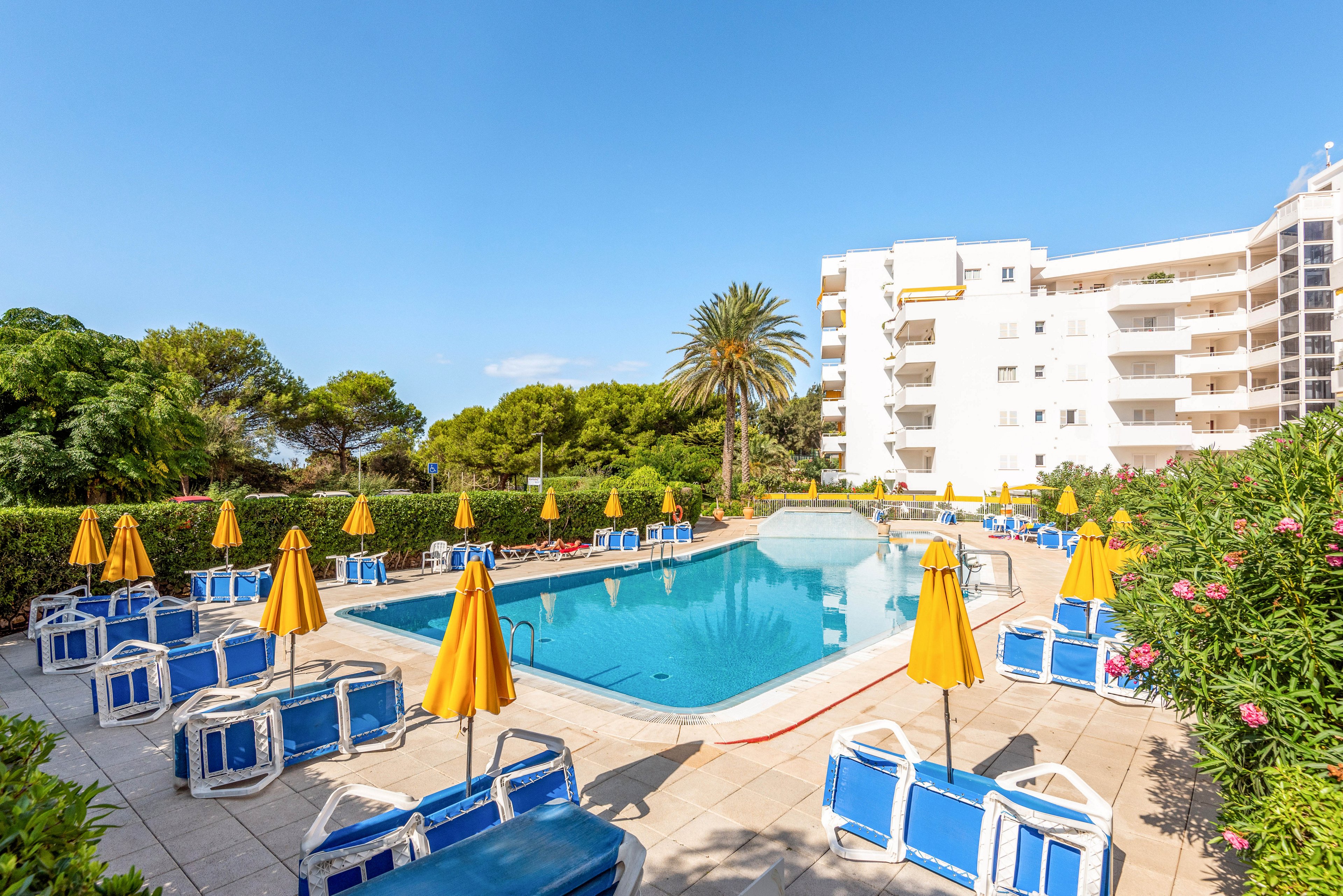 A bright outdoor swimming pool is surrounded by blue lounge chairs and yellow umbrellas, set against a backdrop of lush greenery and a white multi-story hotel building. The scene is bathed in sunlight, creating a cheerful and inviting atmosphere. Palm trees and flowering bushes add to the tropical resort vibe. No people are present, emphasizing a tranquil and relaxing setting. The clear blue sky enhances the overall summery mood.