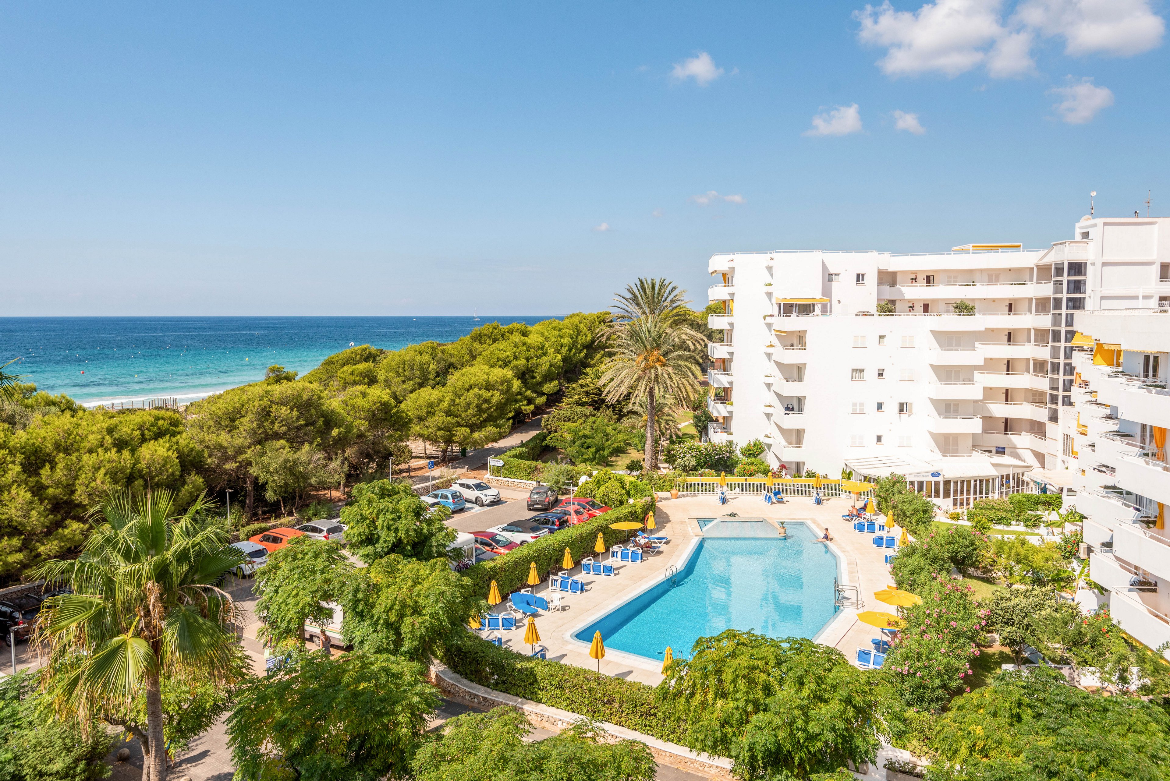 A modern white hotel building overlooks a large outdoor swimming pool surrounded by sun loungers and umbrellas. The setting features lush green trees and gardens, with the blue ocean visible in the background under a clear sky. The scene is bright and inviting, evoking a relaxed vacation atmosphere. No visible text or numbers are present in the image.