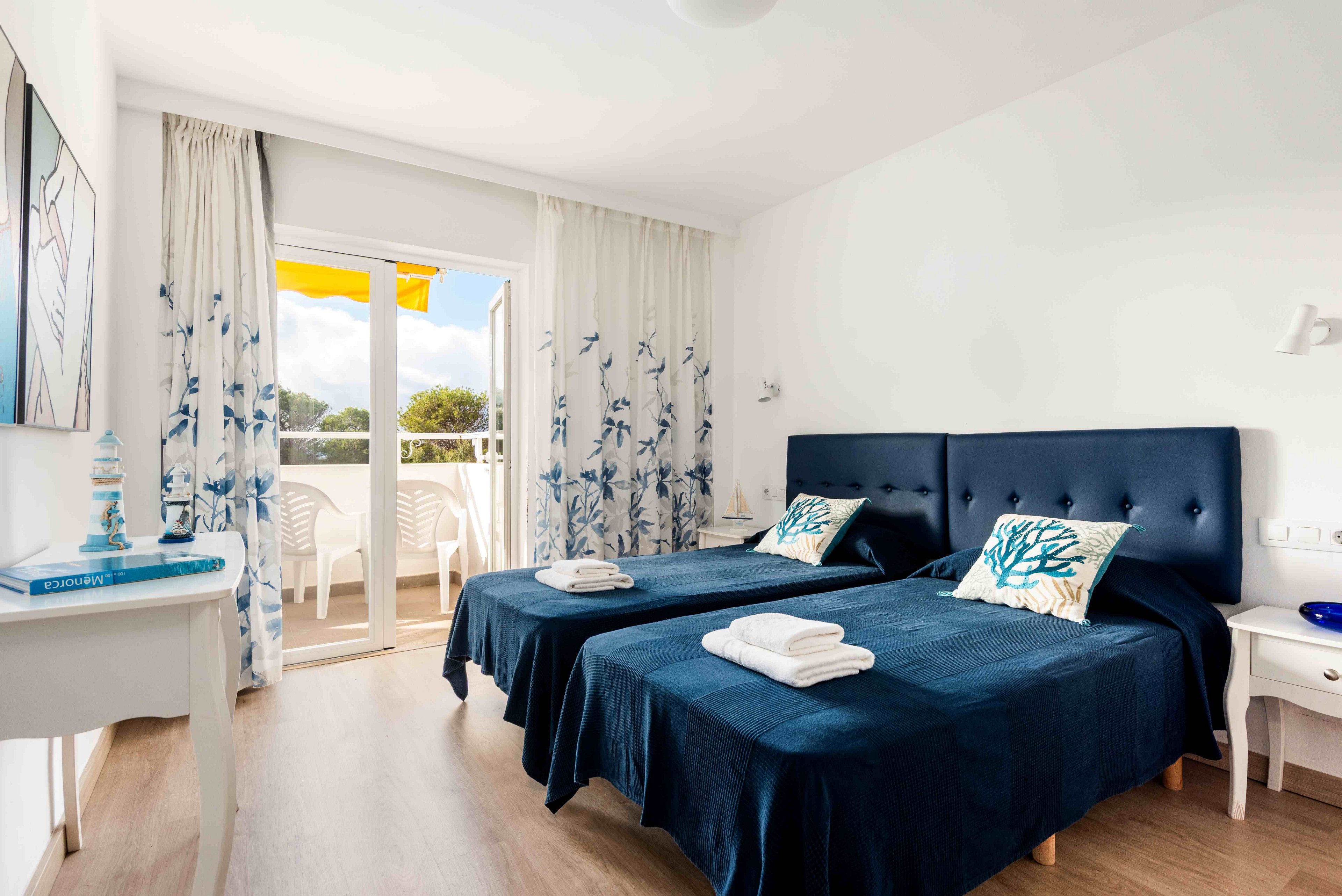 A sunlit hotel room features two twin beds with deep blue bedding and decorative pillows. The space is decorated in a coastal style, with white walls, blue accents, and a balcony visible through glass doors. Natural light fills the room, highlighting the fresh and airy atmosphere. Towels are neatly placed on each bed, and a small table and chair set is visible outside on the balcony.