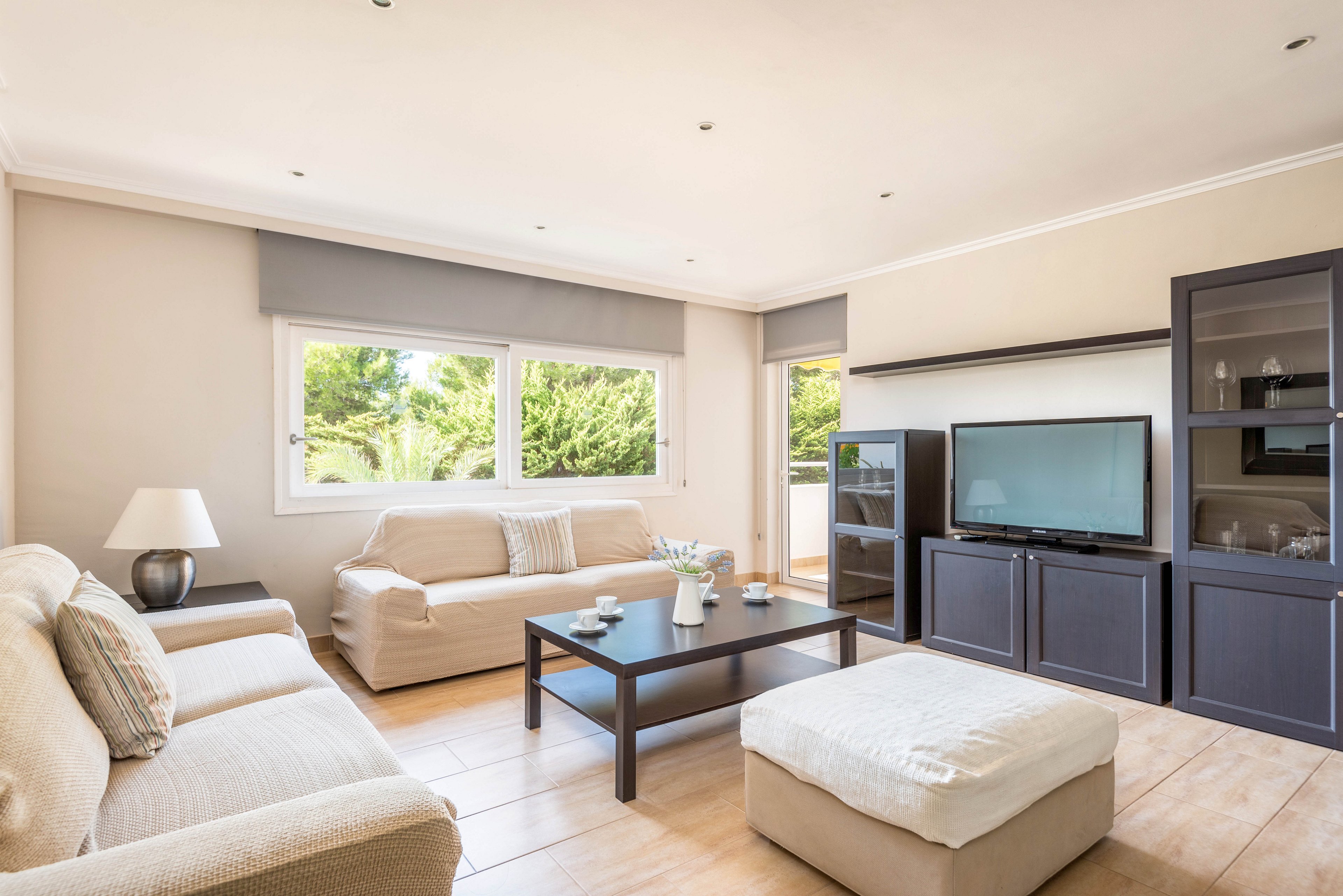 A spacious living room features beige sofas, a large ottoman, and a sleek black coffee table. The setting is well-lit with natural light streaming through wide windows, highlighting the minimalist and contemporary style. The room includes a flat-screen television, shelving units, and subtle decorative accents. The overall palette is neutral with soft beige and gray tones, creating a calm and inviting atmosphere.