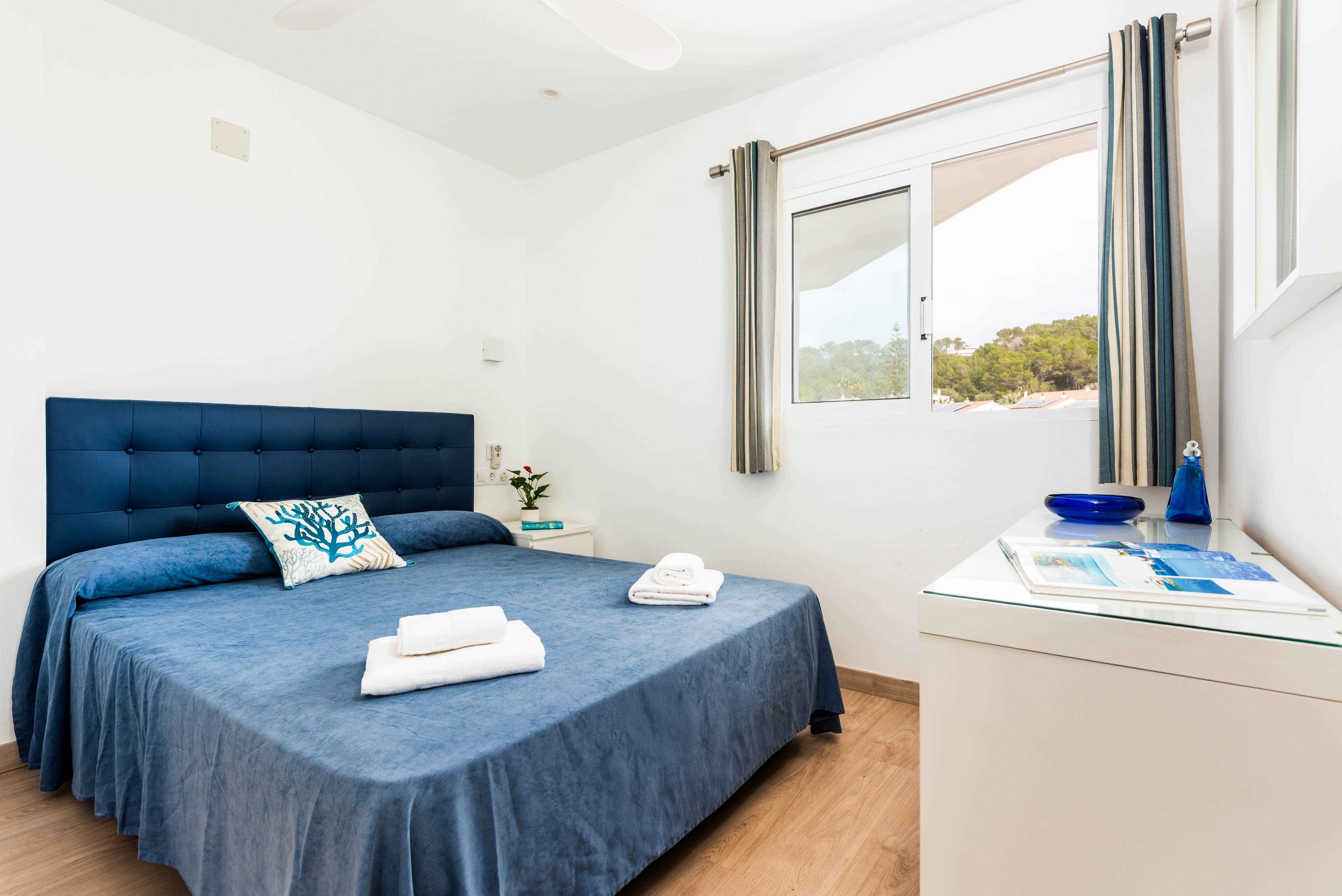 A neatly arranged bedroom features a blue upholstered bed with folded towels and a decorative coral pillow. The room is filled with natural light from a large window, offering a view of greenery outside. Minimalist furnishings include a white dresser with blue accents and a small plant. The overall palette is blue and white, creating a fresh and tranquil atmosphere.