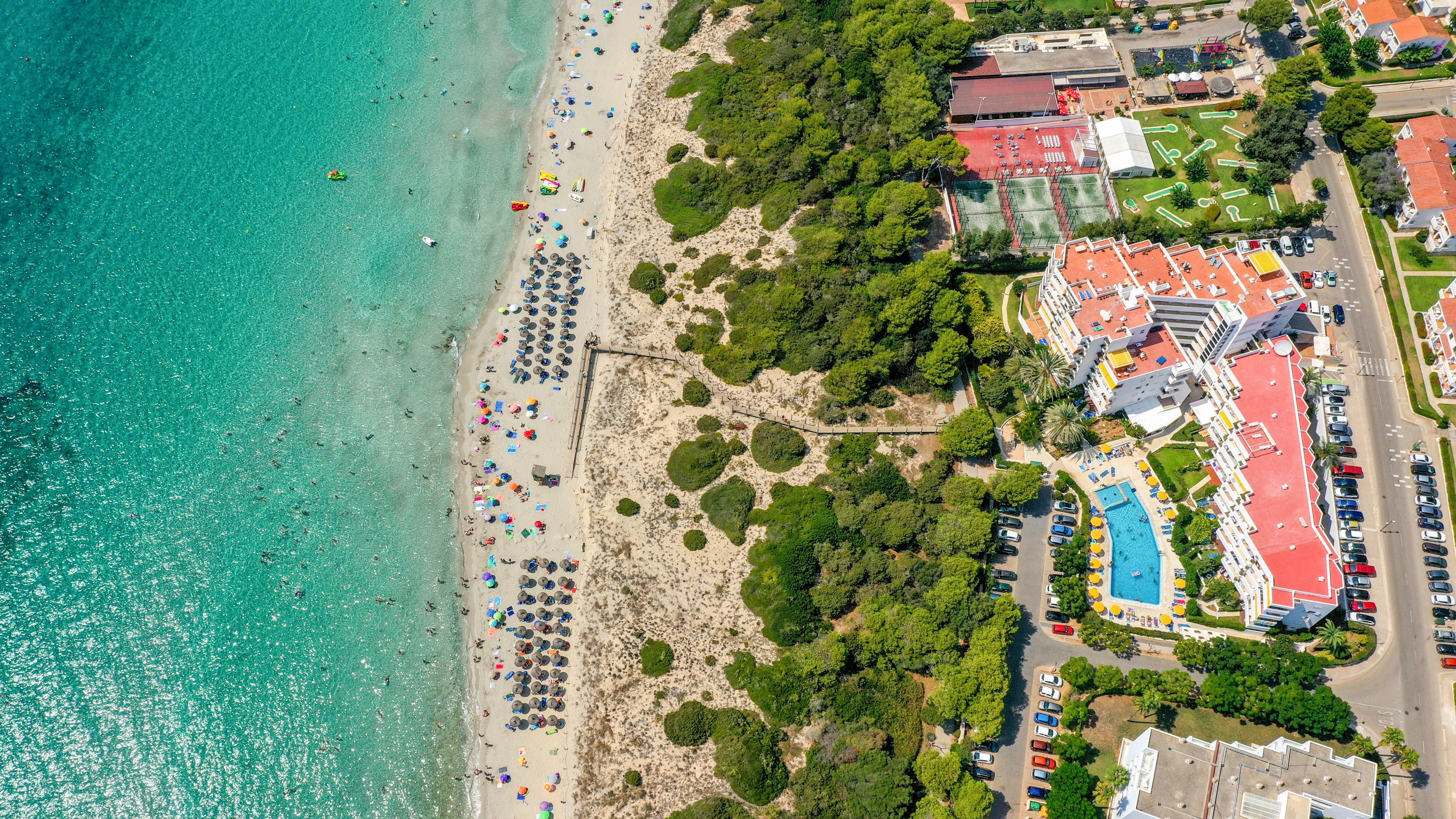 A vibrant aerial photograph captures a beachfront resort adjacent to a sandy coastline with turquoise waters. Numerous sun umbrellas and beachgoers are visible along the shore, while the resort features a large outdoor pool and tennis courts. The scene is lively, with lush greenery separating the resort from the beach, and the overall mood is bright and summery.