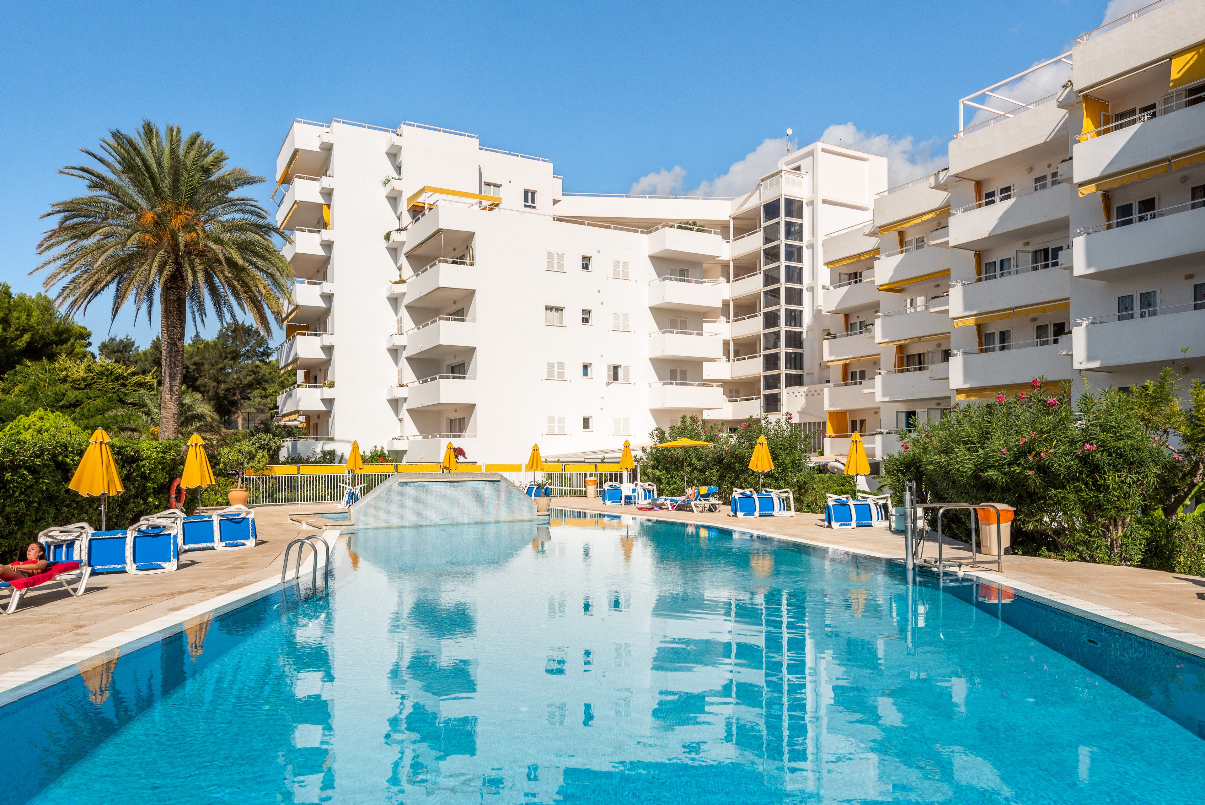 A large outdoor swimming pool is the central focus, surrounded by blue loungers and yellow umbrellas. The setting is a modern hotel or apartment complex with white buildings and balconies, under a clear blue sky. Palm trees and lush greenery add to the vacation atmosphere, creating a bright and inviting mood.