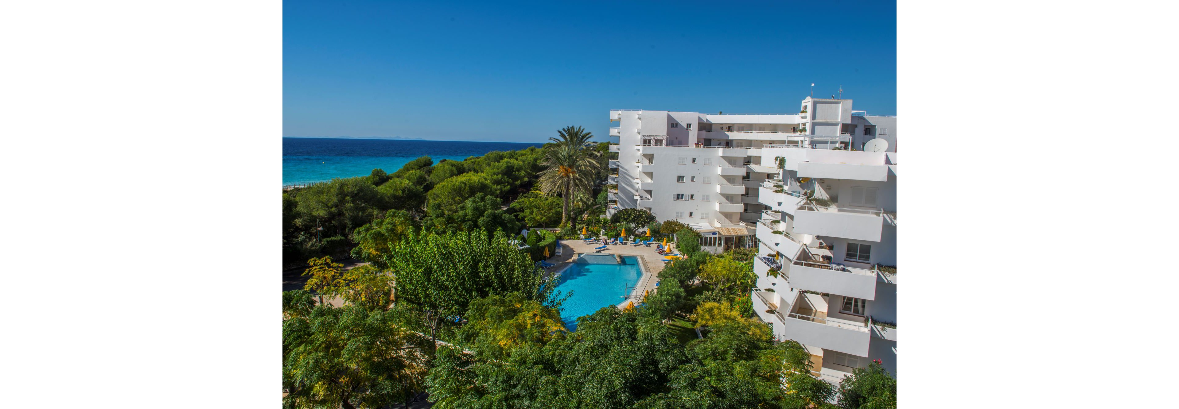 A contemporary white hotel building stands amidst lush greenery, featuring a central outdoor swimming pool. The scene is set on a sunny day with clear blue skies and the ocean visible in the background. Palm trees and landscaped gardens surround the pool area, creating a relaxing vacation atmosphere. No visible text or numbers are present in the image.