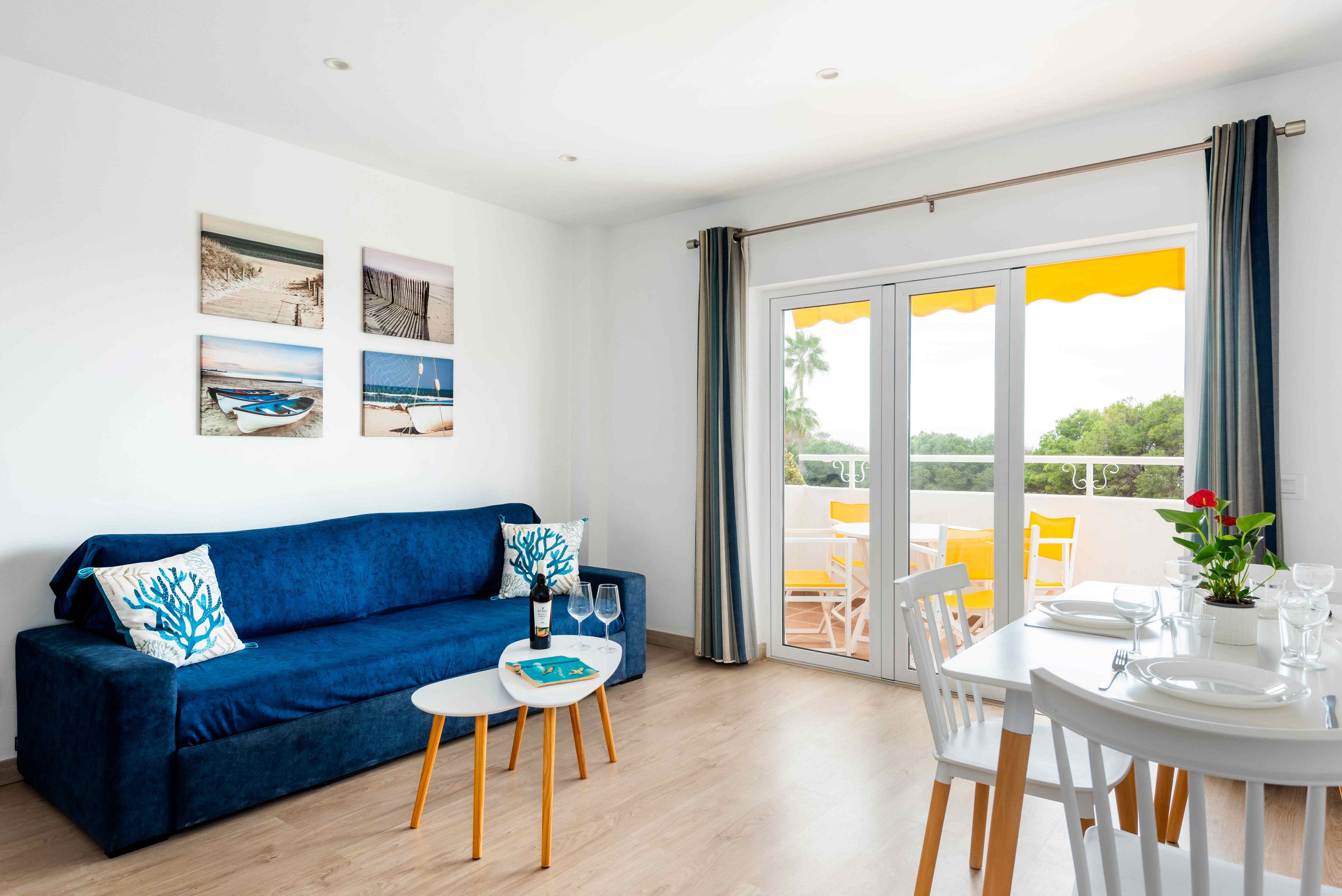 A contemporary living room features a deep blue sofa with decorative pillows and a small coffee table. The space opens to a sunlit balcony with yellow chairs, visible through large glass doors. Wall art with beach scenes and a set dining table add to the inviting atmosphere. The room is styled in a fresh, coastal palette with natural light and minimal decor.