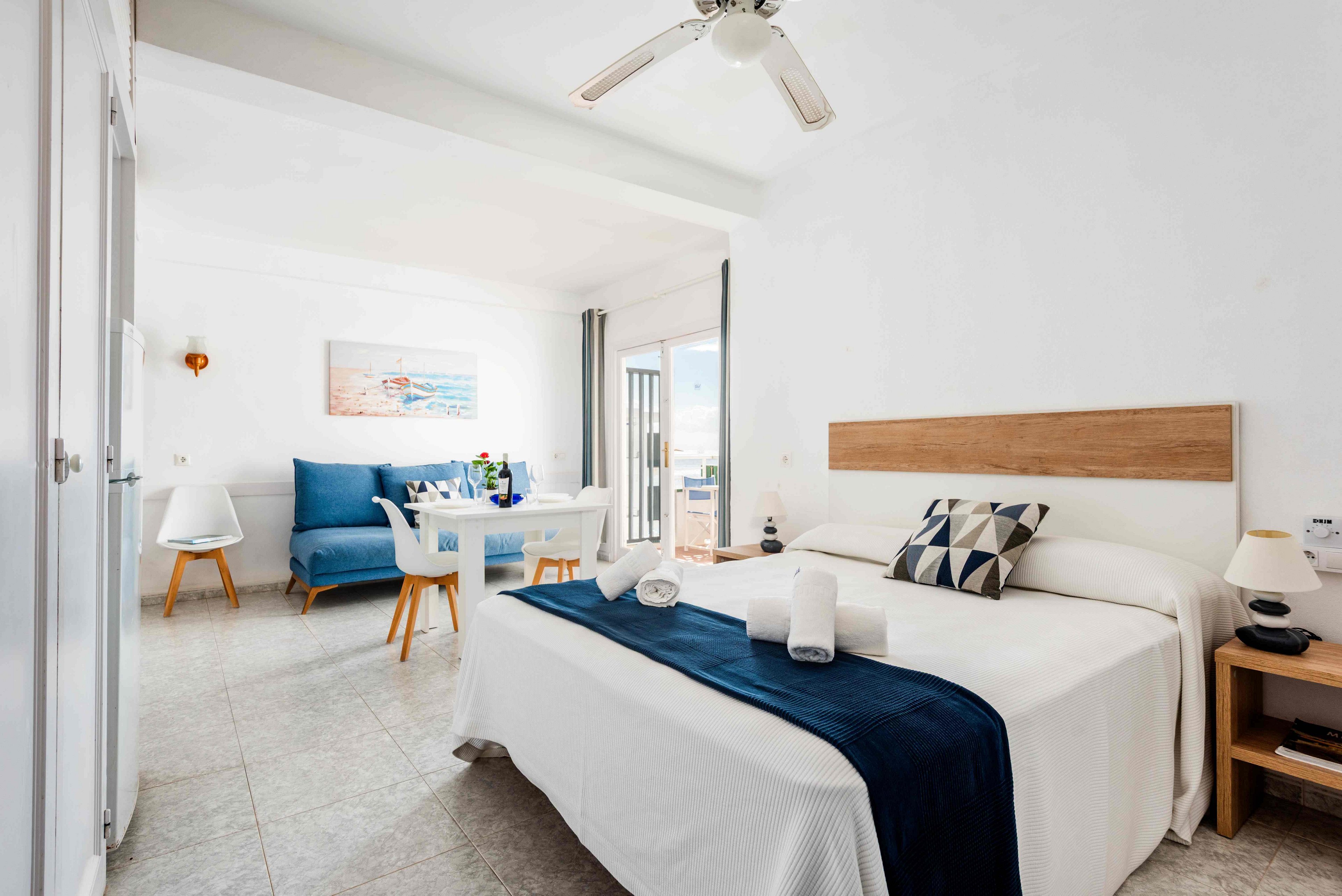 A spacious, sunlit hotel room features a neatly made bed with a navy throw and geometric pillow. The room includes a small dining table, blue sofa, and minimalist decor, with a ceiling fan above and a door leading to a balcony. The palette is dominated by white and blue tones, creating a fresh and inviting atmosphere. Rolled towels are placed on the bed, and a painting of a beach scene hangs on the wall. The overall style is contemporary and clean.