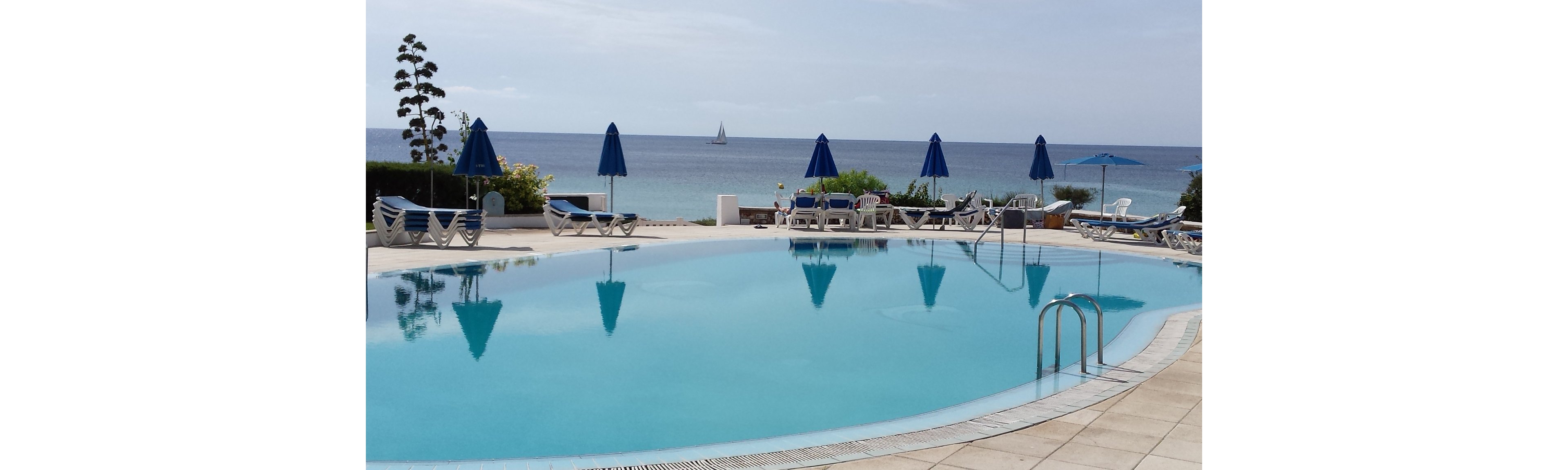 A tranquil swimming pool is set beside the ocean, with several blue umbrellas and lounge chairs arranged around the pool deck. The scene overlooks calm blue water and a distant sailboat, evoking a peaceful, relaxing atmosphere. The pool area is empty, emphasizing the quiet and inviting mood under a clear sky.