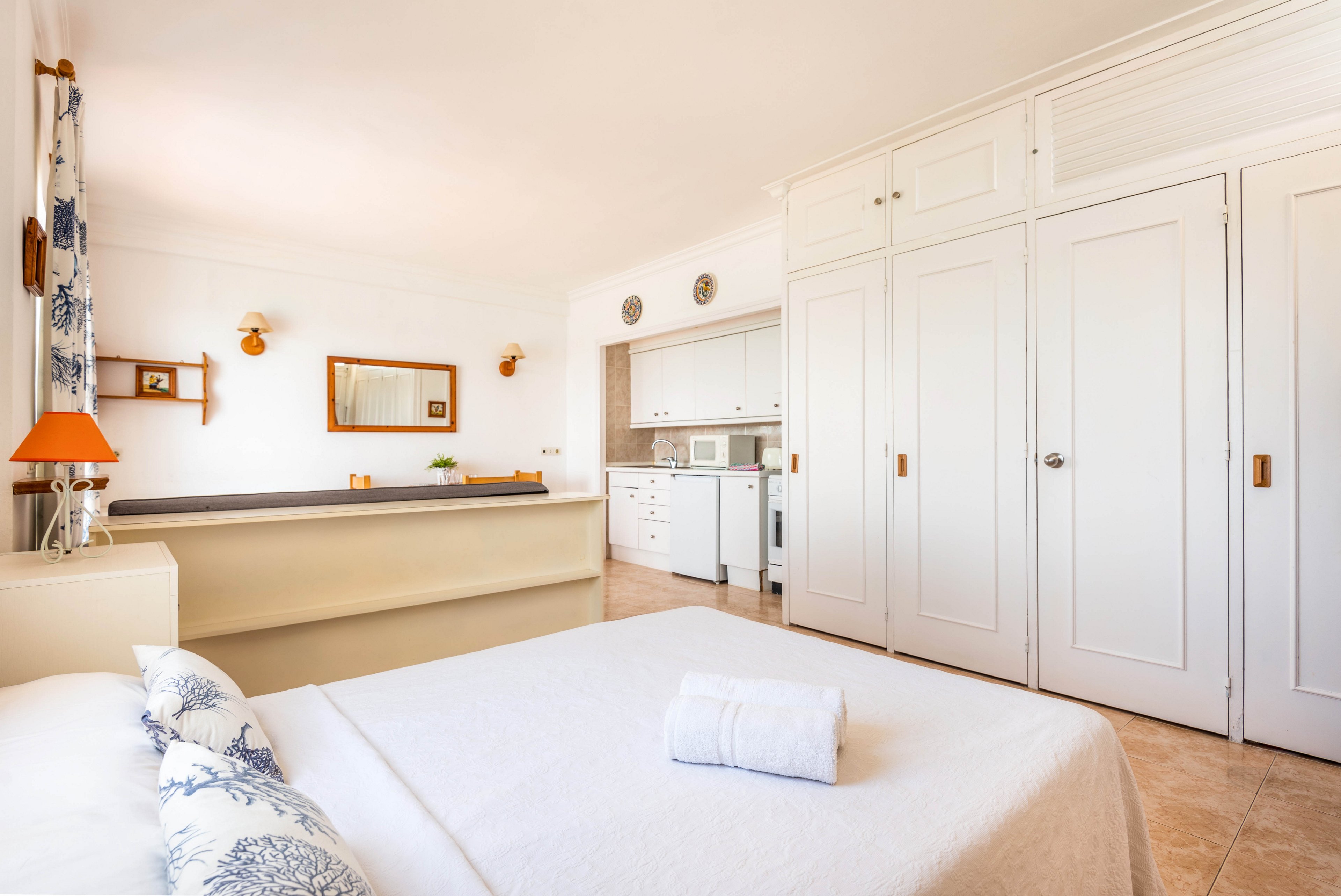 A spacious, sunlit bedroom features a neatly made bed with white linens and blue patterned pillows. The room opens into a small kitchen area, separated by a low partition, and is decorated with white cabinetry and light wood accents. The overall palette is neutral with touches of blue and orange, creating a calm and inviting atmosphere. No visible people or explicit numeric values are present.