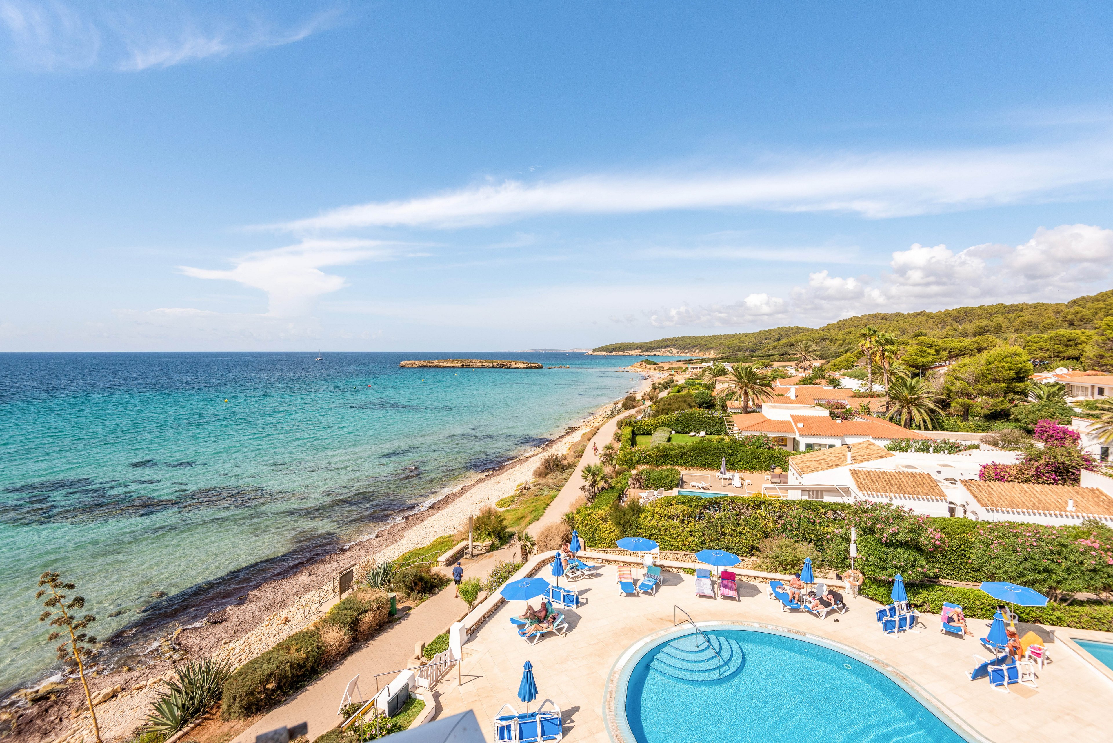 A scenic resort pool area is shown with sun loungers and blue umbrellas, set beside a clear turquoise sea. The image captures a relaxed vacation atmosphere with people sunbathing and swimming. Mediterranean-style villas with terracotta roofs line the coast, surrounded by lush greenery. The sky is mostly clear, enhancing the vibrant blue tones of the water and pool.