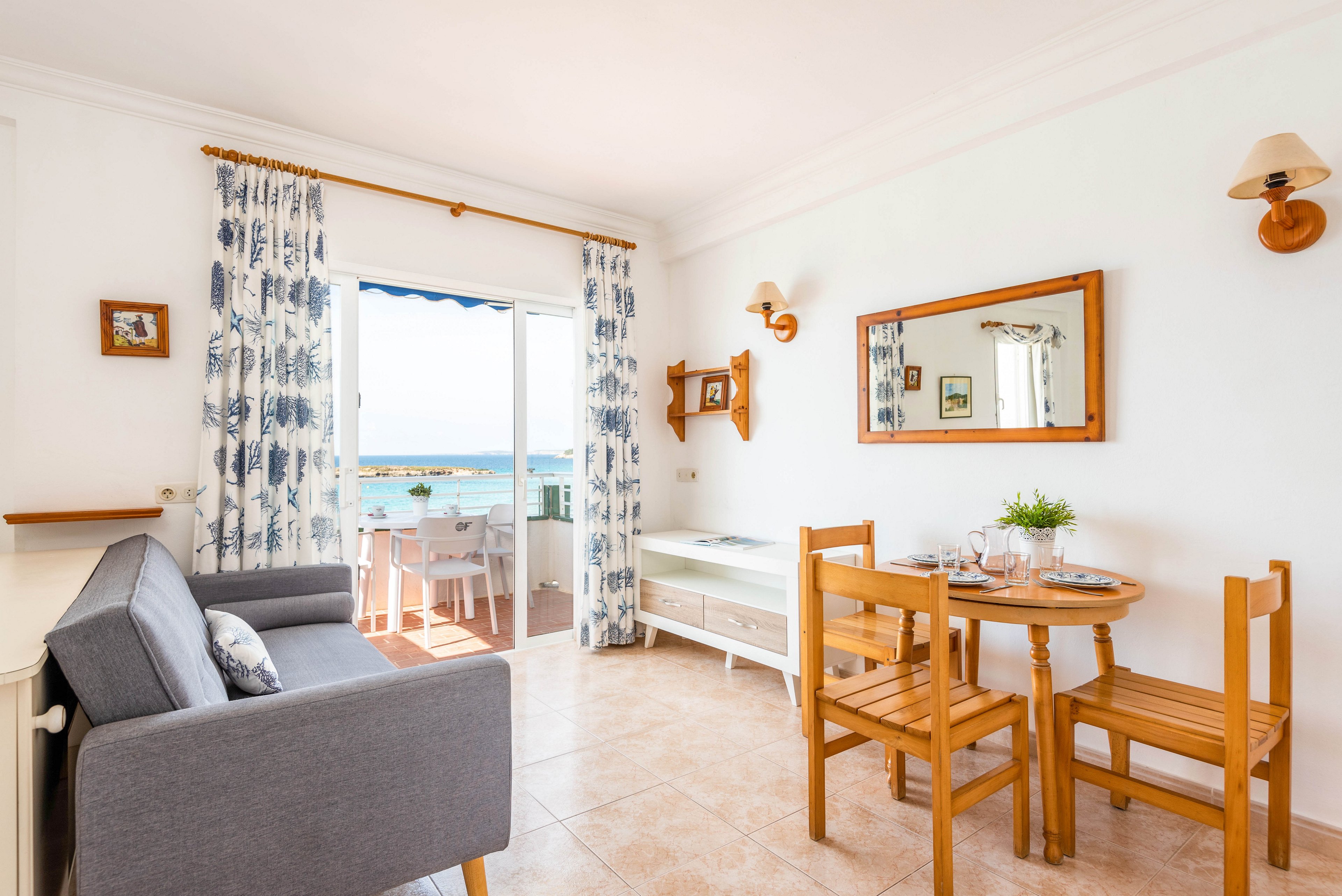 A sunlit living room features a gray sofa, wooden dining table, and chairs, with a large mirror on the wall. The space opens to a balcony overlooking the ocean, framed by patterned curtains. The decor is minimal and airy, with white walls and natural wood accents, creating a relaxed, inviting atmosphere.