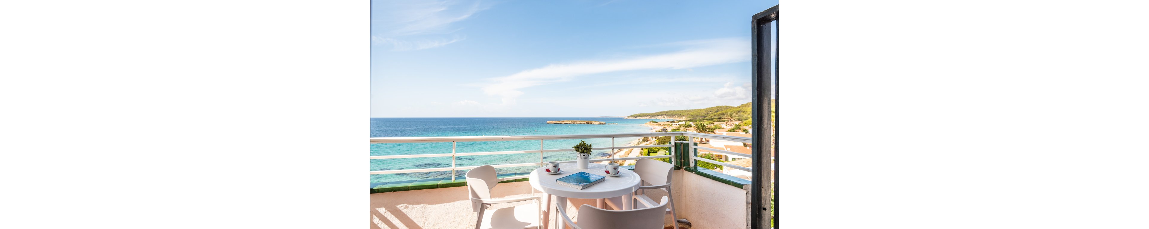 A bright outdoor balcony features a round white table with four chairs, set against a stunning turquoise ocean backdrop. The table holds two coffee cups, a small potted plant, and a blue magazine, suggesting a relaxing morning scene. The clear sky and sunlight create a cheerful, inviting atmosphere, perfect for leisure or vacation. The coastal landscape and open railing enhance the sense of openness and tranquility.