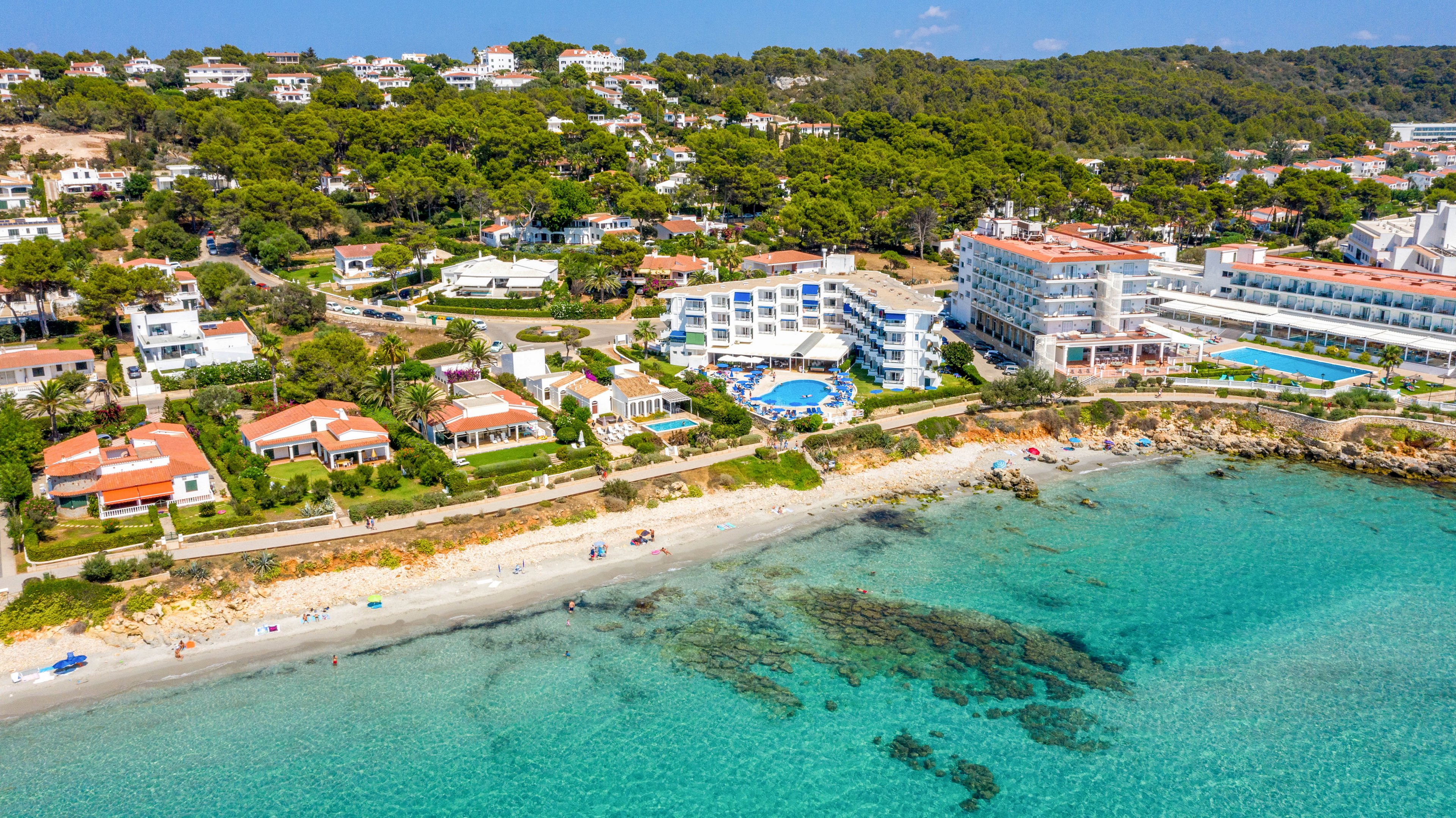 This image captures a scenic aerial view of a coastal resort area with white modern hotels and villas lining a sandy beach. The turquoise water is clear, revealing underwater rocks near the shore, while guests enjoy the beach and pool. The setting is lush with greenery and forested hills in the background, creating a tranquil vacation atmosphere. No visible text or numbers are present in the image.