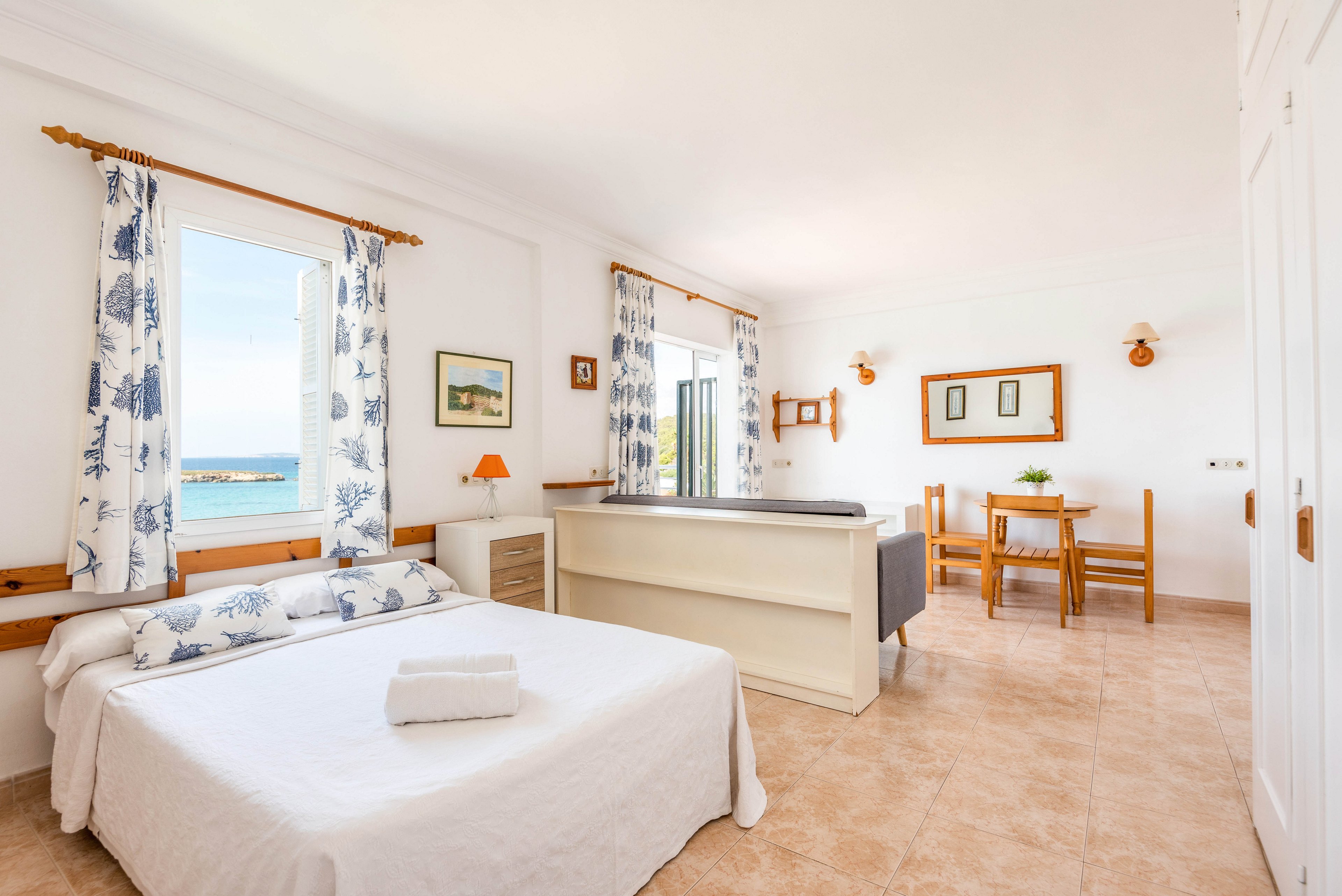 A sunlit bedroom features a neatly made double bed with folded towels, set against a backdrop of large windows overlooking the ocean. The room is decorated in a fresh, coastal style with blue and white curtains, light wood furniture, and minimalistic decor. A small dining table and chairs are positioned near the window, and the overall palette is airy and inviting. The space is uncluttered, with tiled floors and white walls enhancing the natural light.