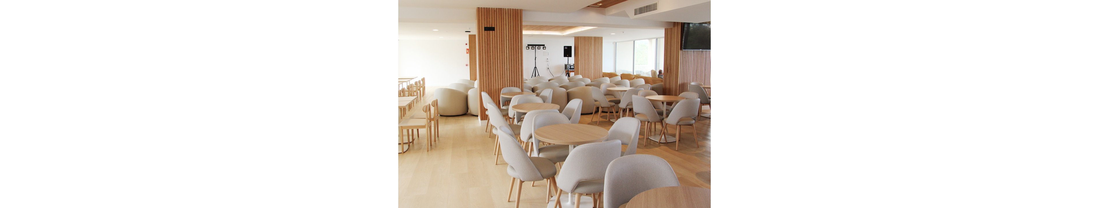 The image shows a spacious cafeteria featuring round tables and cushioned chairs in neutral tones. The setting is bright with natural light, wooden panel accents, and minimalist decor. No people are present, and the mood is calm and inviting. The palette consists of beige, light wood, and white, creating a contemporary and clean atmosphere.