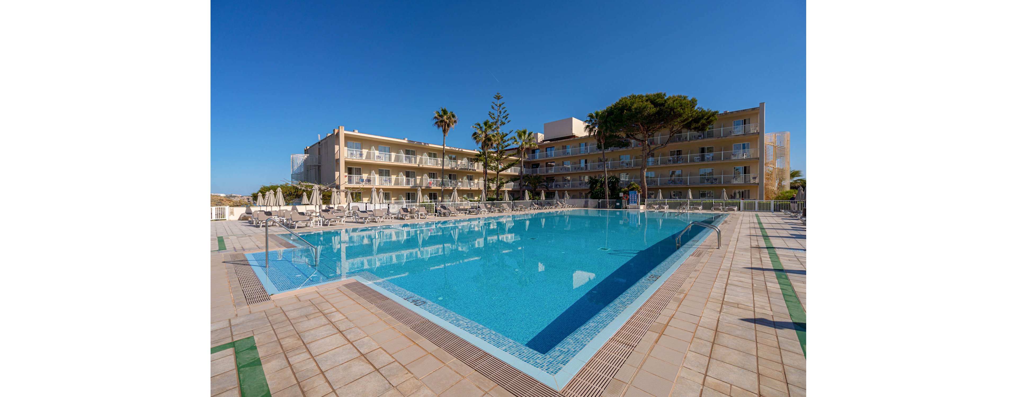 A large rectangular swimming pool is the central feature of this outdoor hotel area, surrounded by sun loungers and umbrellas. The hotel buildings are visible in the background, with balconies overlooking the pool. The setting is bright and sunny, with clear blue skies and palm trees adding to the vacation atmosphere. No visible text or numeric values are present in the image.