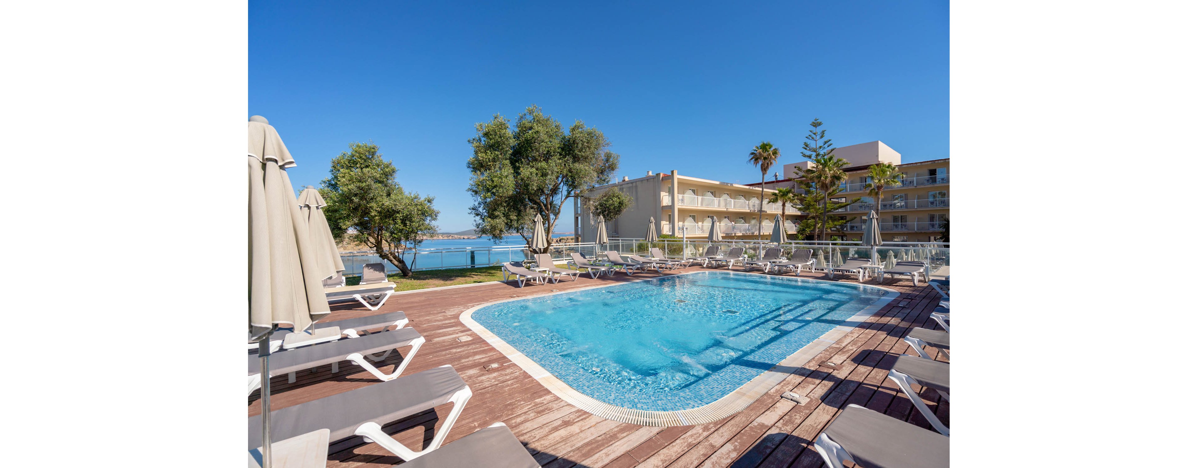 The image shows a clear blue swimming pool surrounded by numerous sun loungers and umbrellas on a wooden deck. The setting is an outdoor hotel area with buildings and palm trees visible in the background, overlooking a scenic waterfront. The atmosphere is bright and sunny, with a vibrant blue sky and tranquil mood. No visible text or numbers are present in the image.