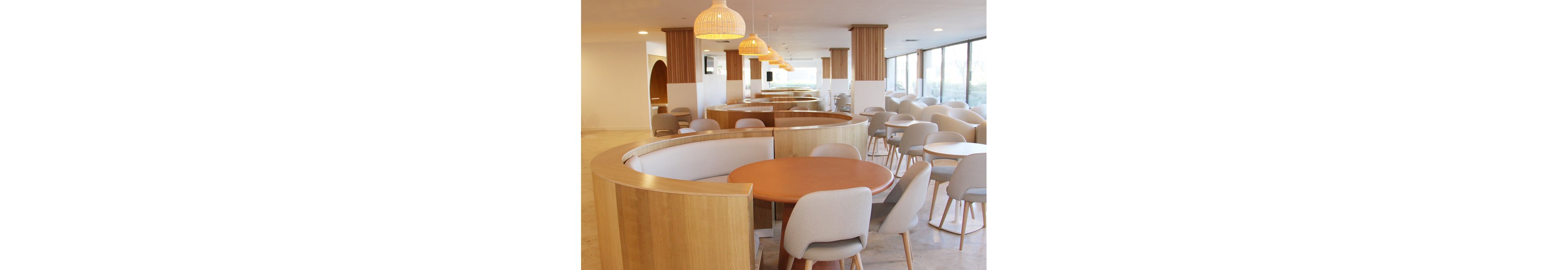 A spacious cafeteria features contemporary curved wooden booths and light-colored chairs arranged around round tables. Pendant lights with woven shades hang from the ceiling, casting a warm glow over the neutral-toned interior. Several people are seated in the background, engaging in conversation. Large windows allow natural light to fill the space, enhancing the airy and inviting atmosphere.