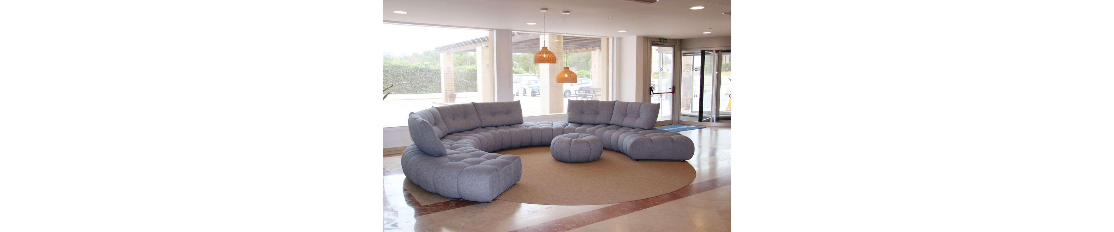 A large, light gray curved sectional sofa with matching ottoman is arranged on a round beige rug in a bright, open lobby. The space features floor-to-ceiling windows, pendant lights, and marble flooring. The overall mood is contemporary and inviting, with natural light enhancing the neutral color palette.