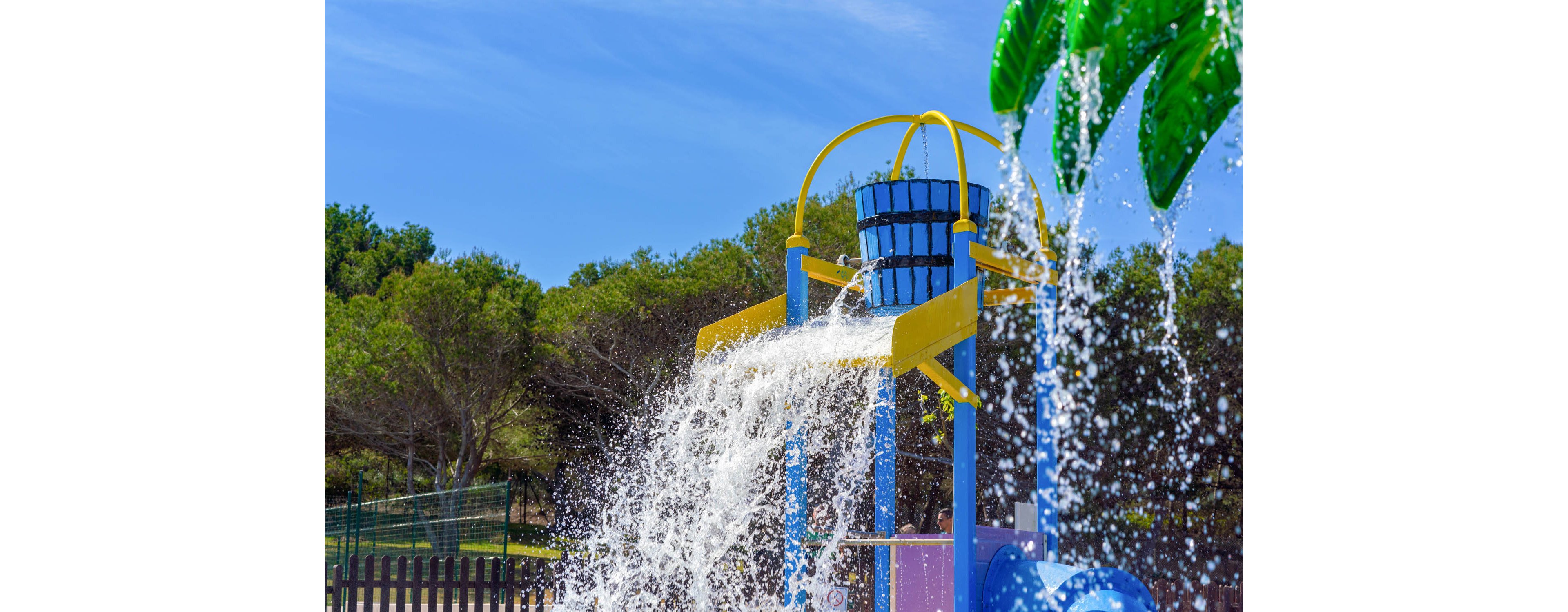 A large blue and yellow tipping bucket pours water onto a play area in an outdoor splash park. The scene is set under a clear blue sky, surrounded by green trees and fencing. Water cascades dramatically, creating a lively and refreshing atmosphere, with palm tree decorations visible. No visible text or numeric values are present in the image.