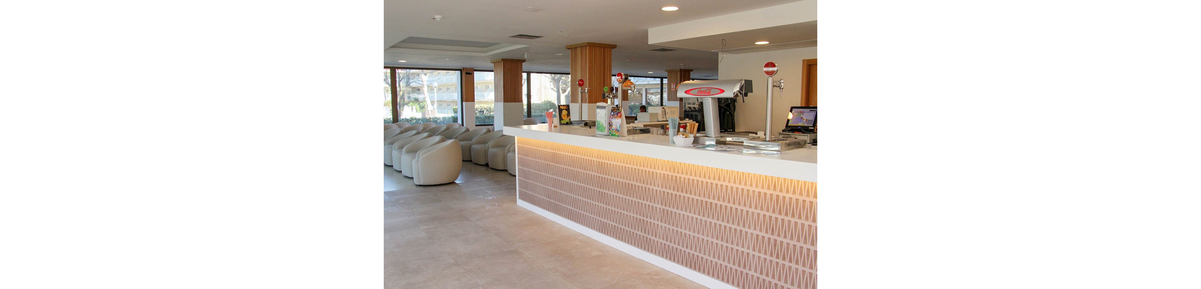 A contemporary hotel lobby bar features a sleek white counter with geometric tile patterns and a visible beverage dispenser. The spacious setting includes rows of cushioned chairs and large windows providing natural light. The atmosphere is clean and inviting, with neutral tones and minimalist decor. The bar area displays a sign with the text 'Amstel' clearly visible.